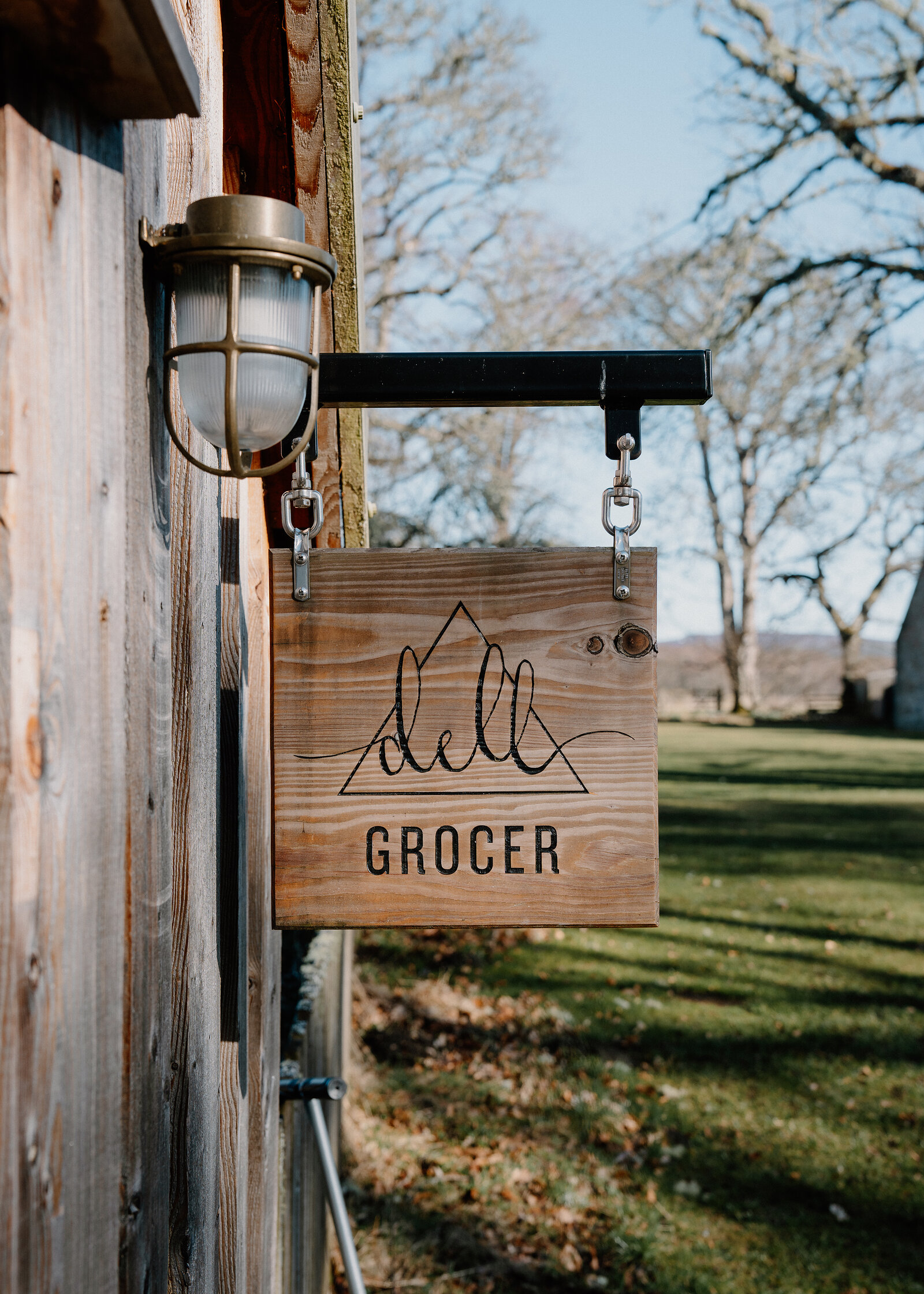 The Dell Grocer's carved wooden sign hanging from a bracket on a weathered timber wall, the lawn beyond.