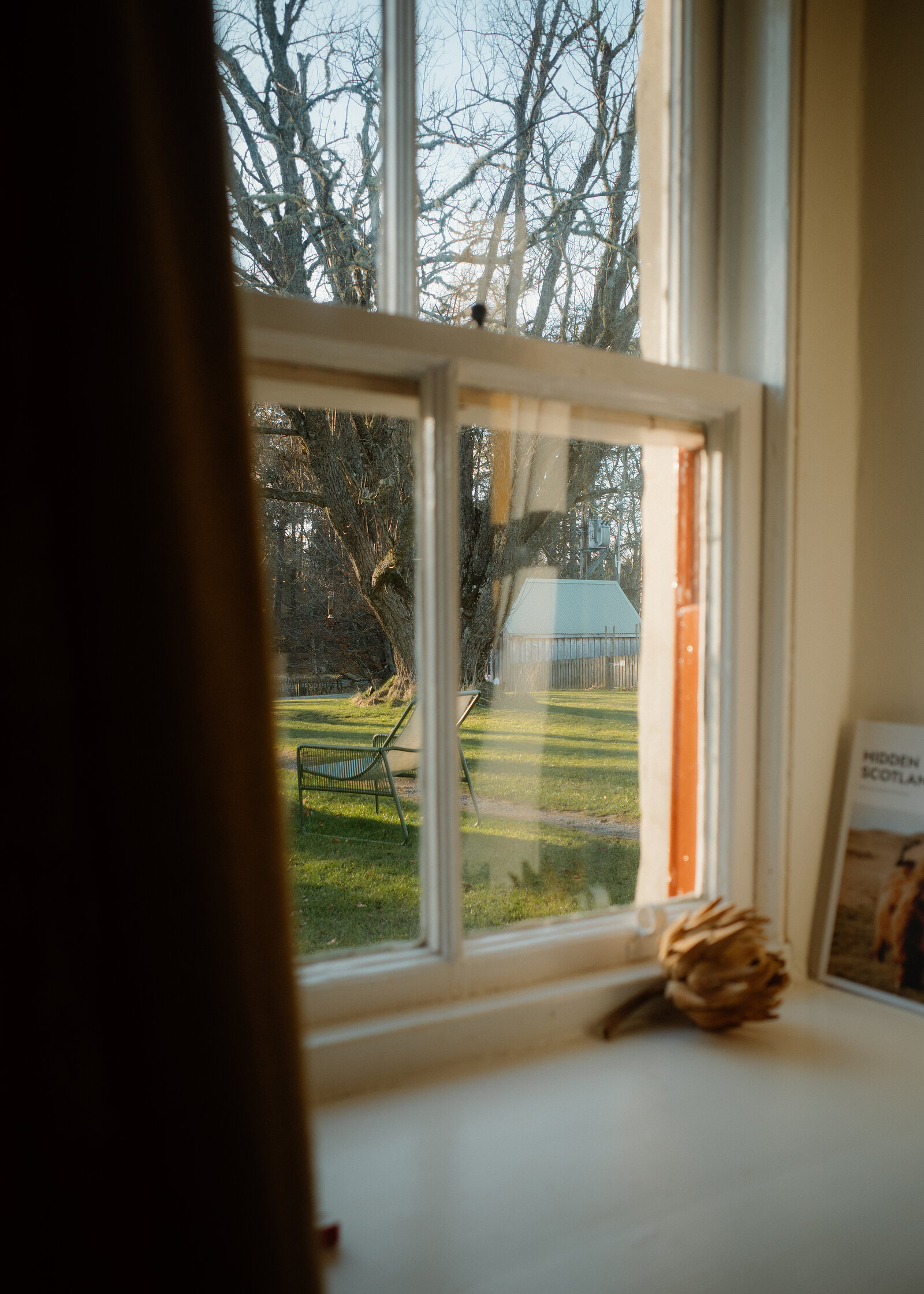 A sash window looking out onto the lawn and the trees, a curtain drawn back.