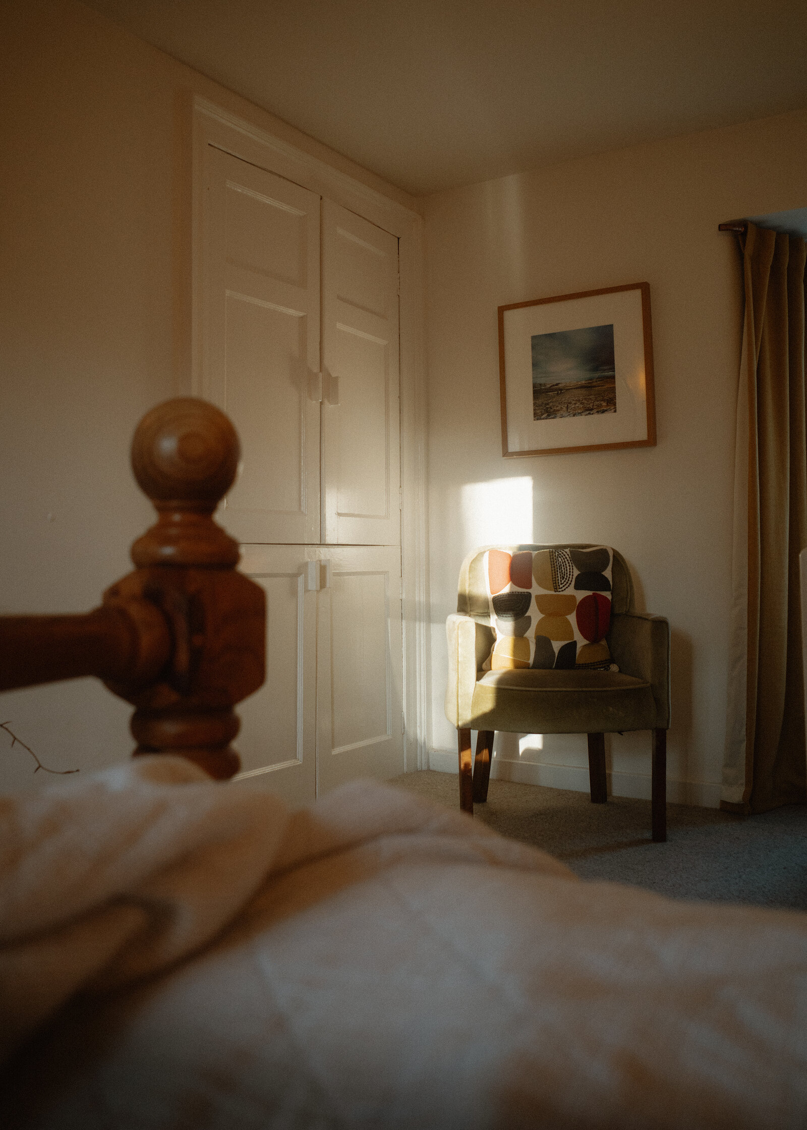 West Dell's bedroom from the bed's pine post, an armchair by the window and mustard curtains.
