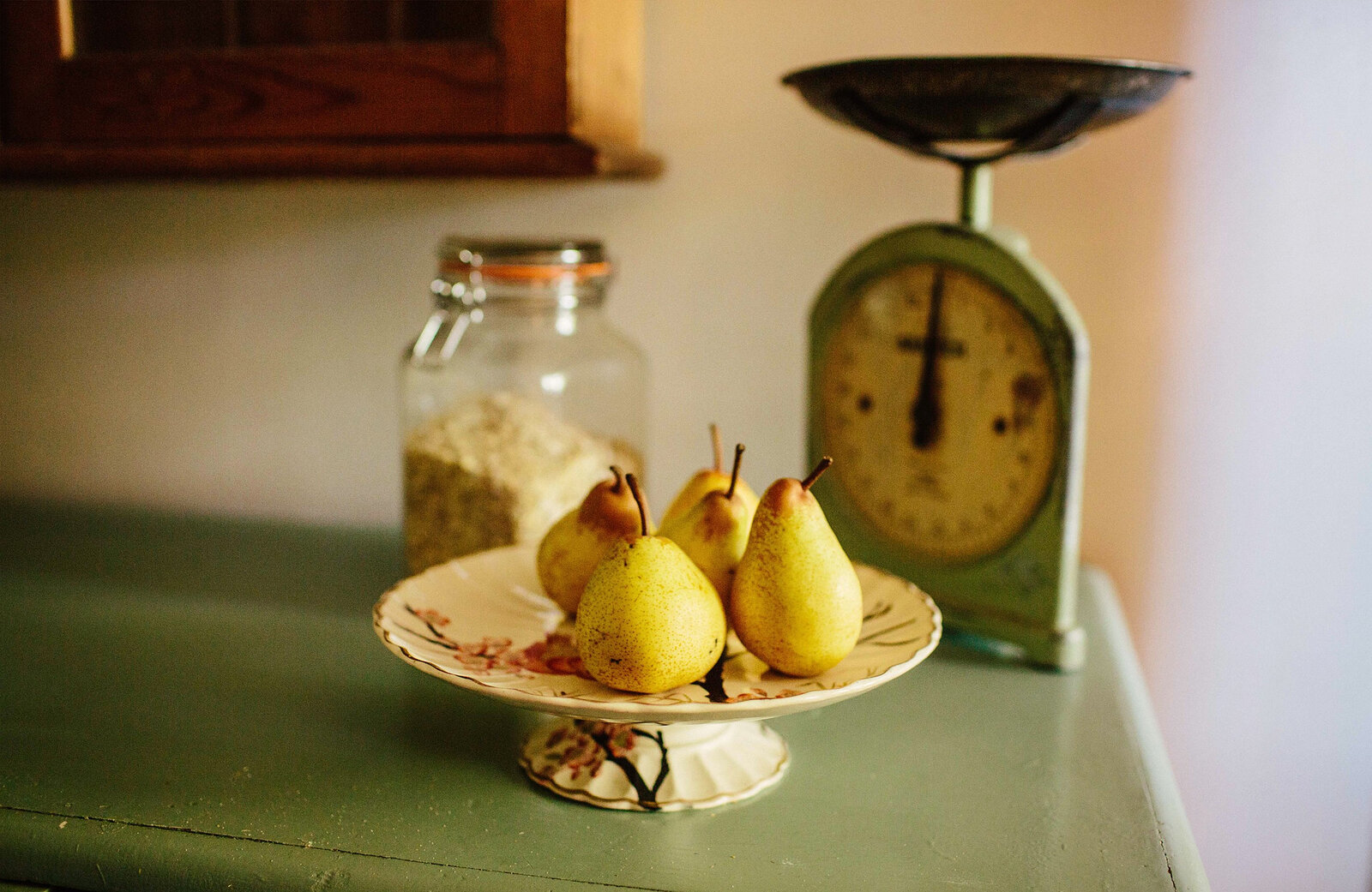 Pears in a dish beside a pair of old green kitchen scales and a jar of oats.