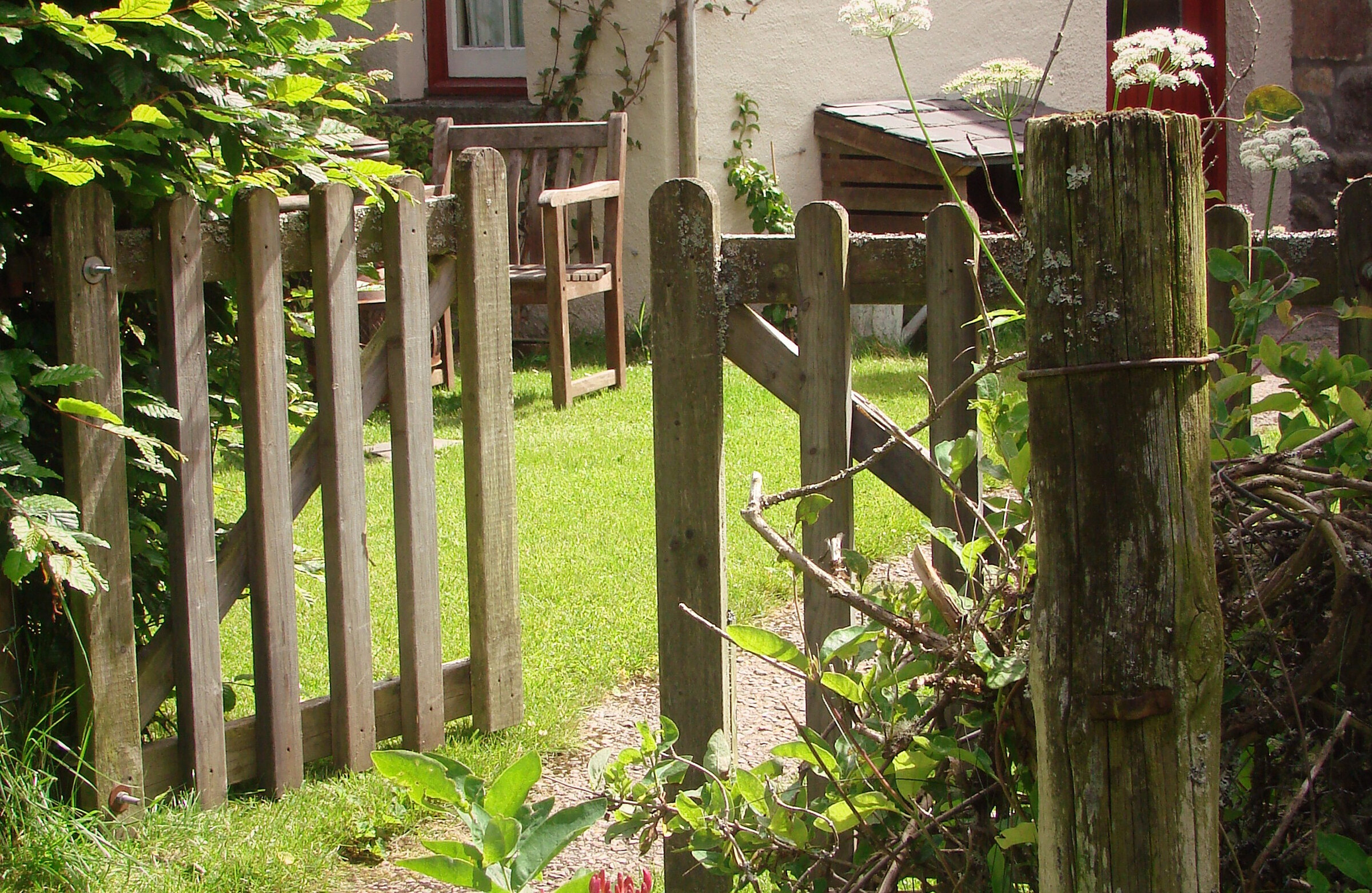 A wooden gate in a picket fence opens onto Little Dell's green garden, with cow parsley and a wooden bench beyond.