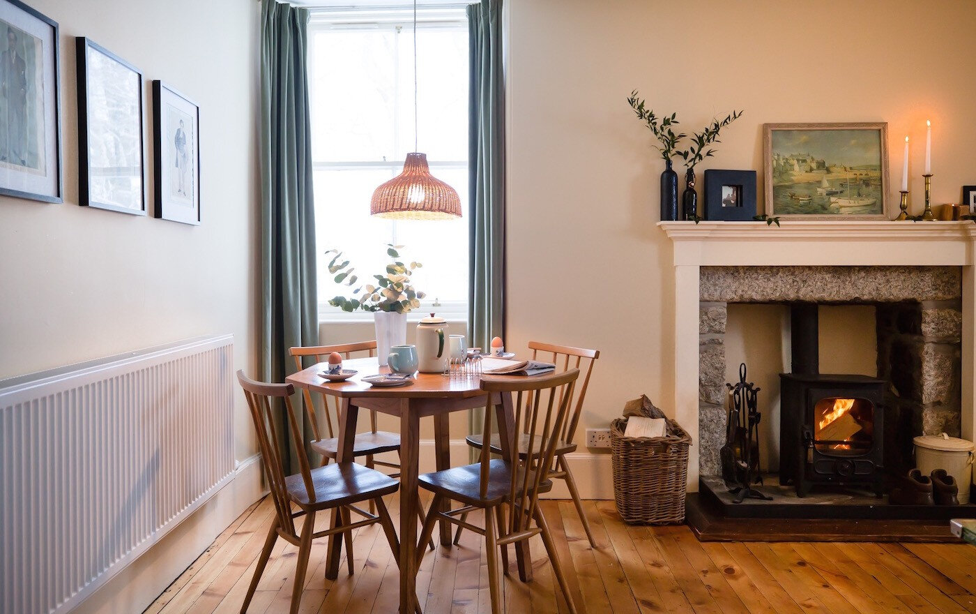 A small round dining table by the tall window, with the stove burning in the fireplace across the room.