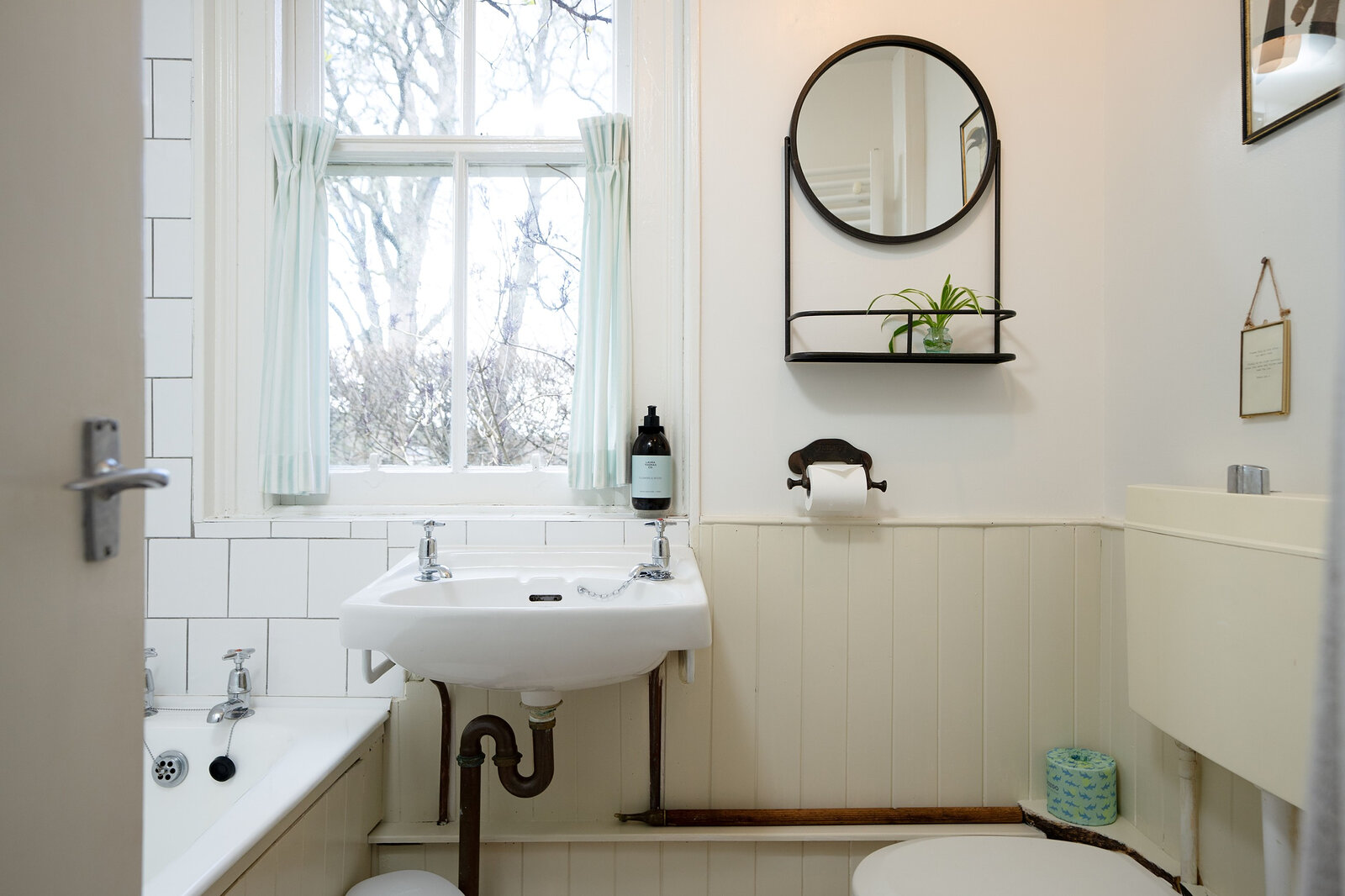 A white bathroom with a bath, a small basin and a round mirror beneath a window onto the trees.