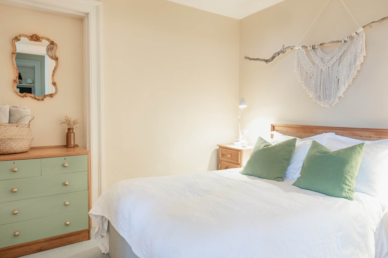 The double bedroom with white linen and green cushions, a macramé hanging and a gilt mirror over a green chest.