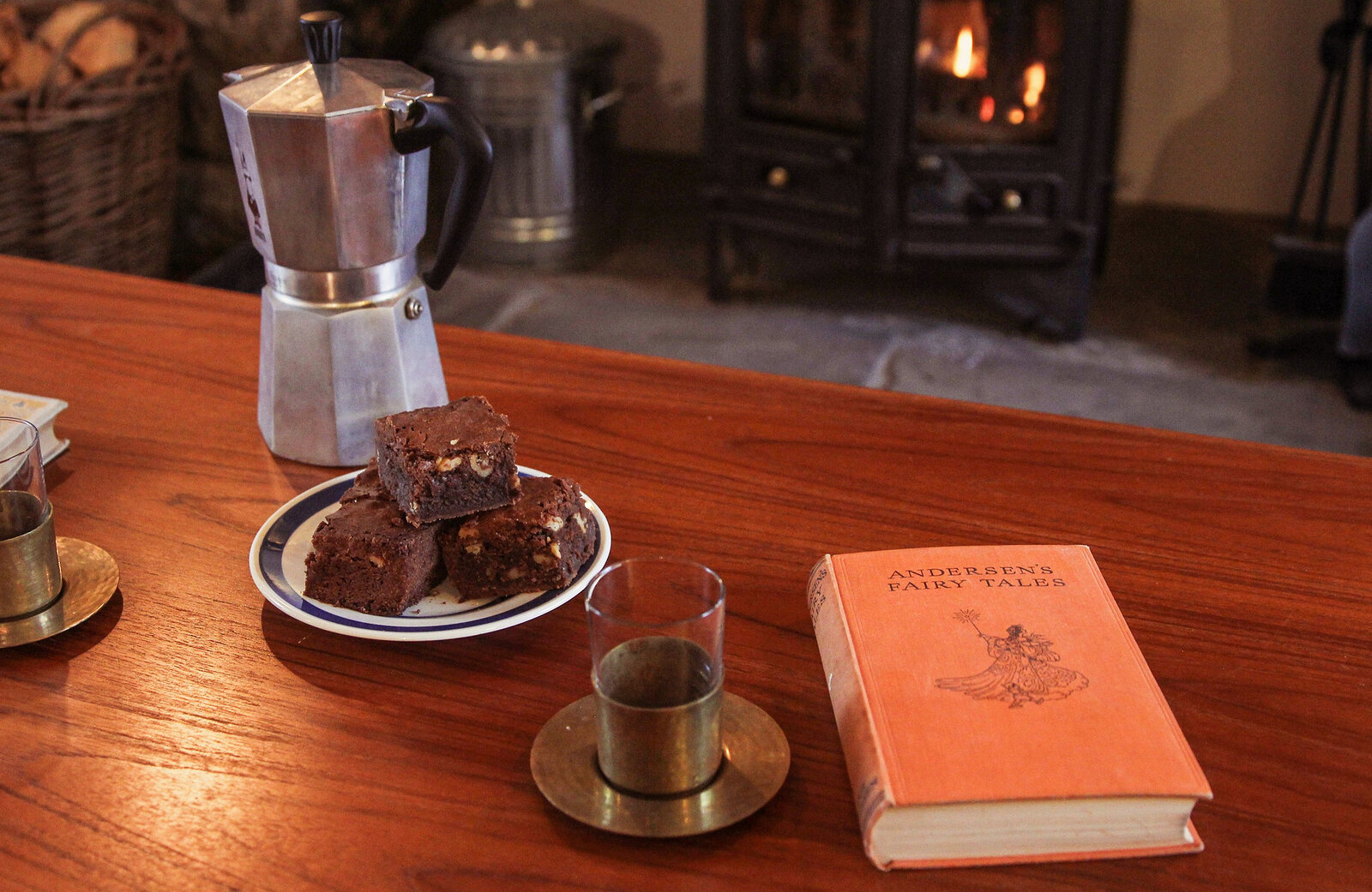 Brownies and a coffee pot on the table, a book beside them and the stove glowing behind.