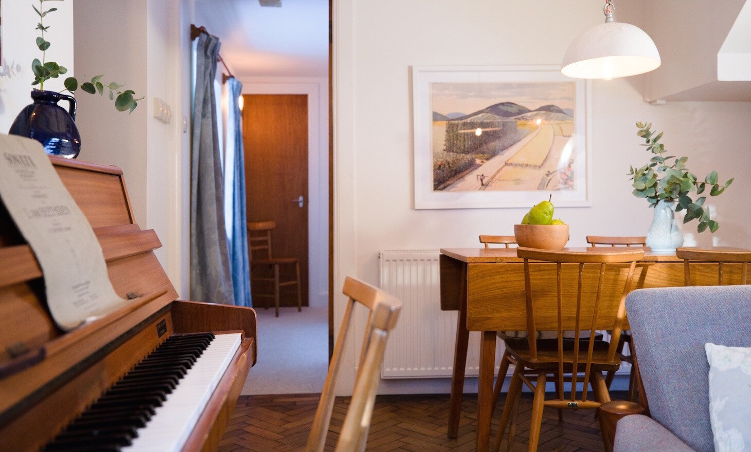 The piano beside the dining table, with a print of a beach on the wall and a door through to the hall.