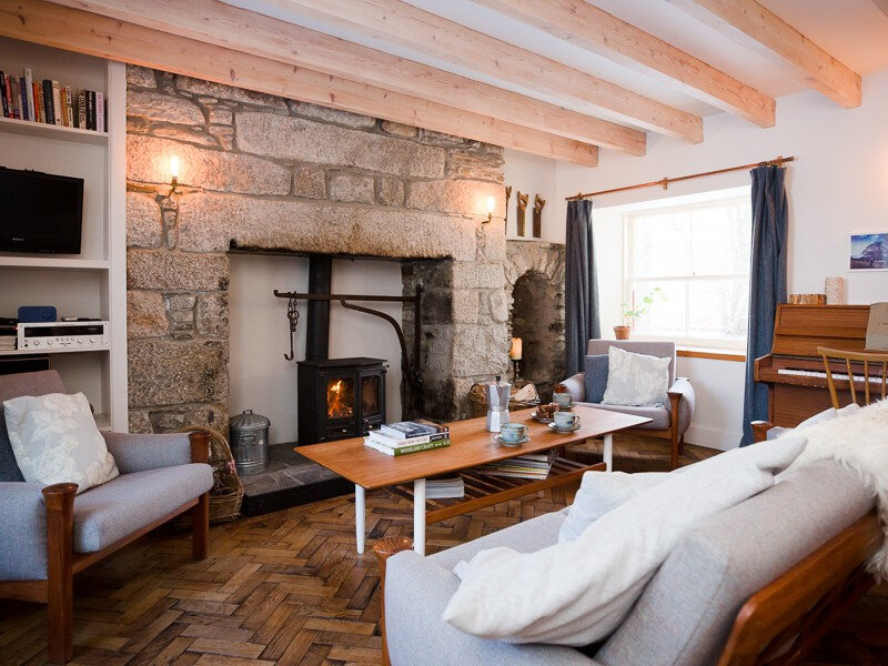 The sitting room under its beams: the granite range surround and stove, a coffee table, sofas, bookshelves and the piano.