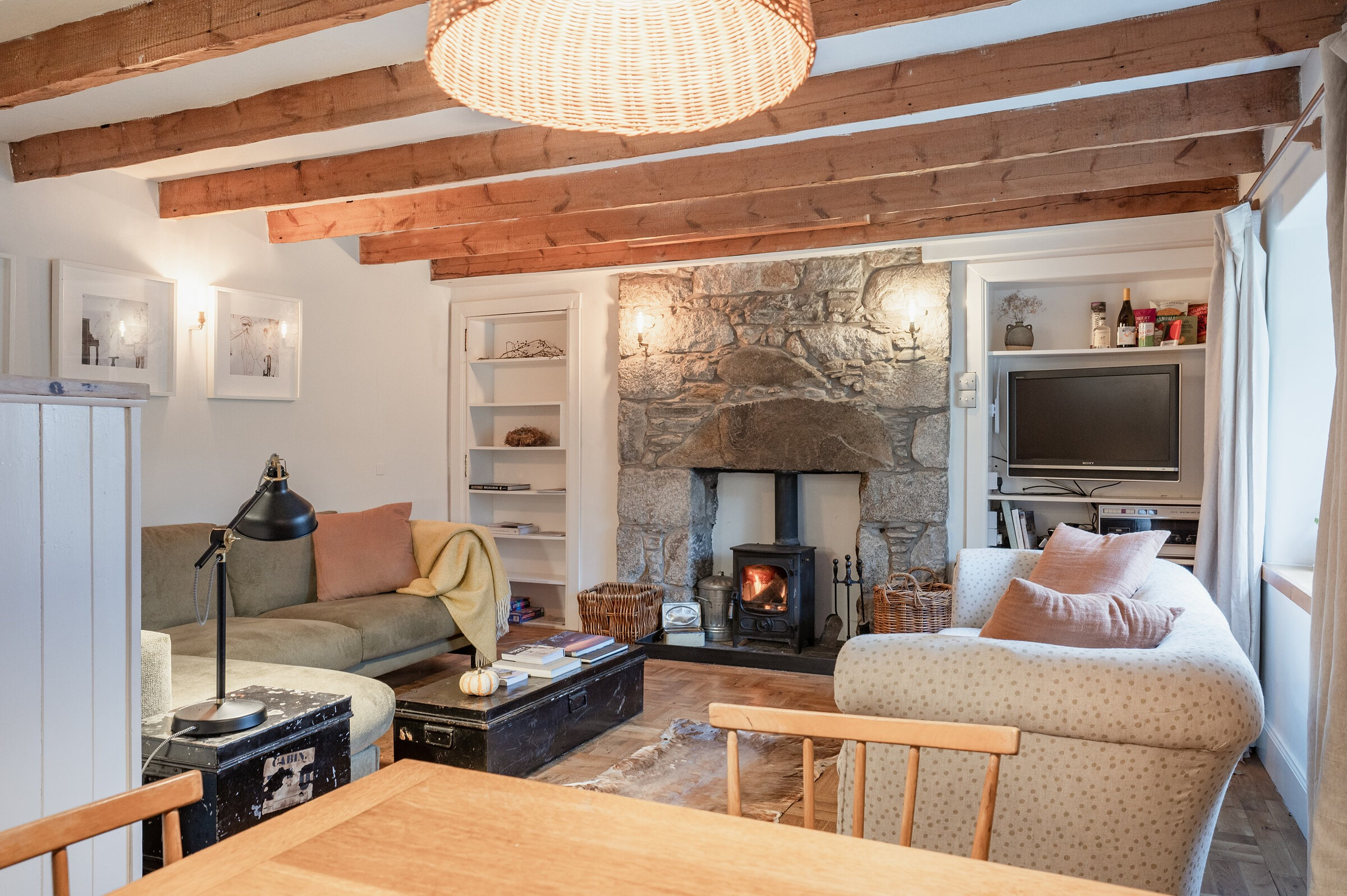 The open sitting room under timber beams, a stove lit in the rough granite fireplace, with sofas and a dining table.