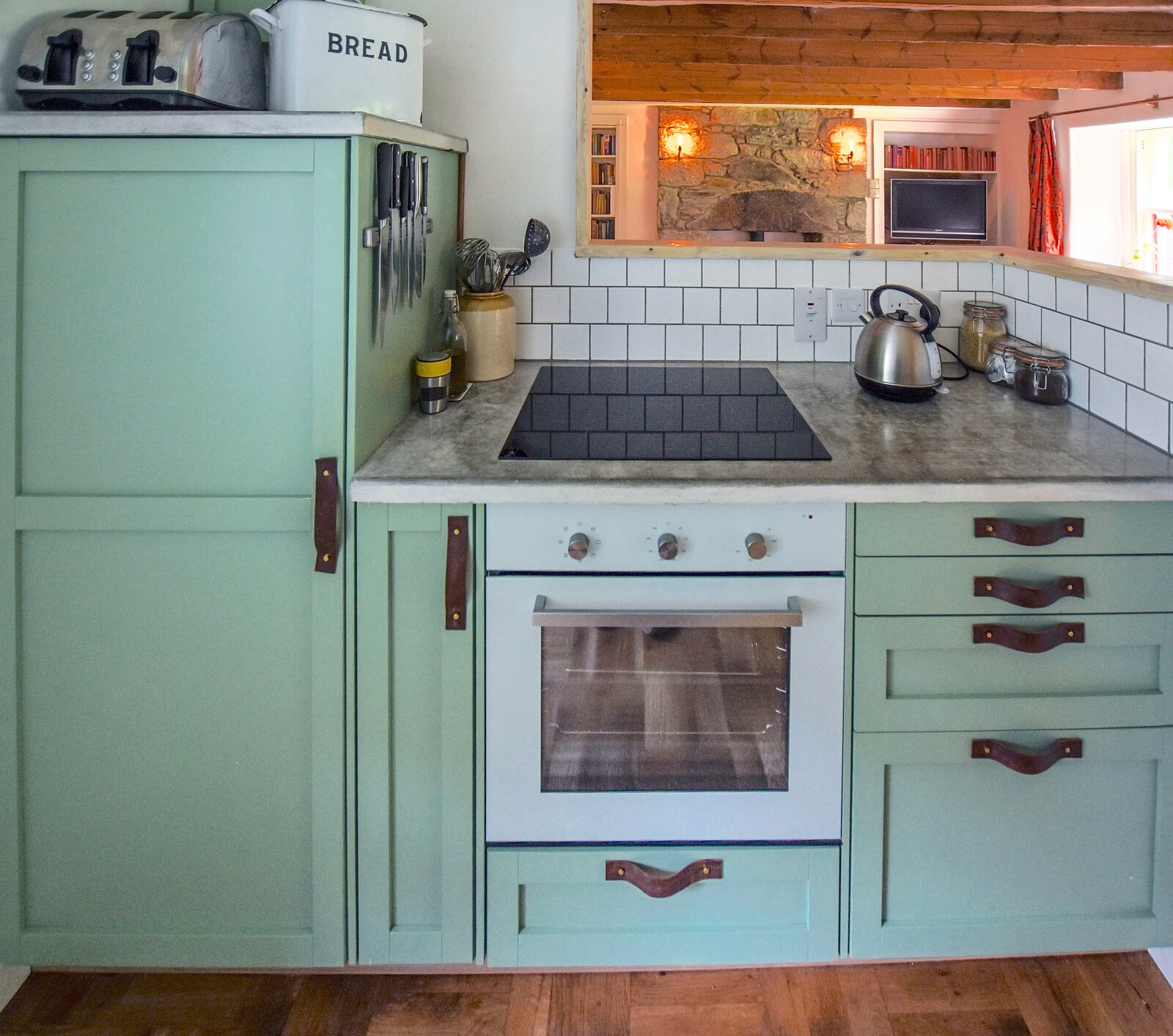 The kitchen with sage-green units and leather handles, a white tiled wall and a window at the end.