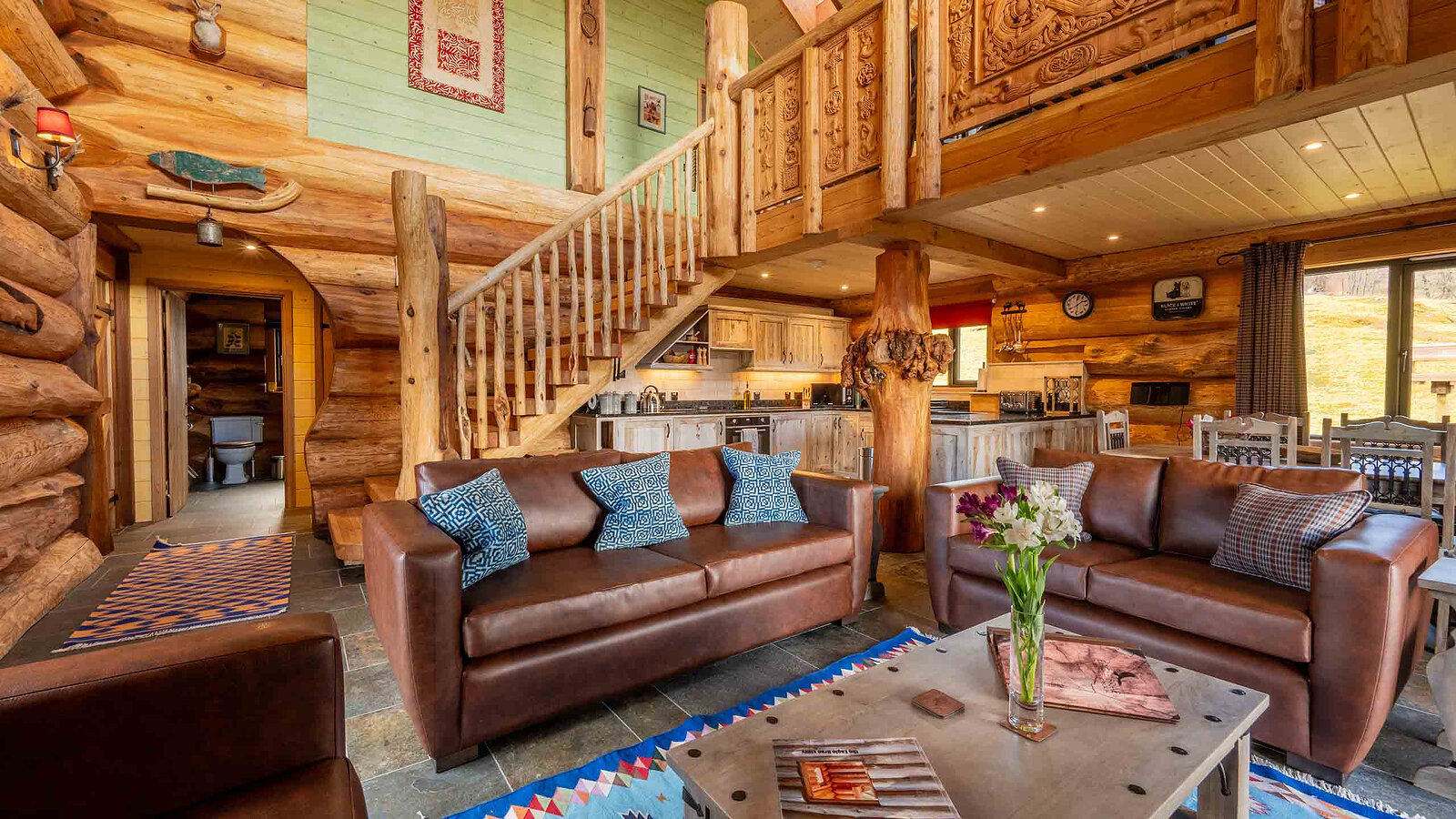 Aquila's open living room: leather sofas on a Himalayan rug, the stair and carved gallery rising to the mezzanine, the kitchen beyond.