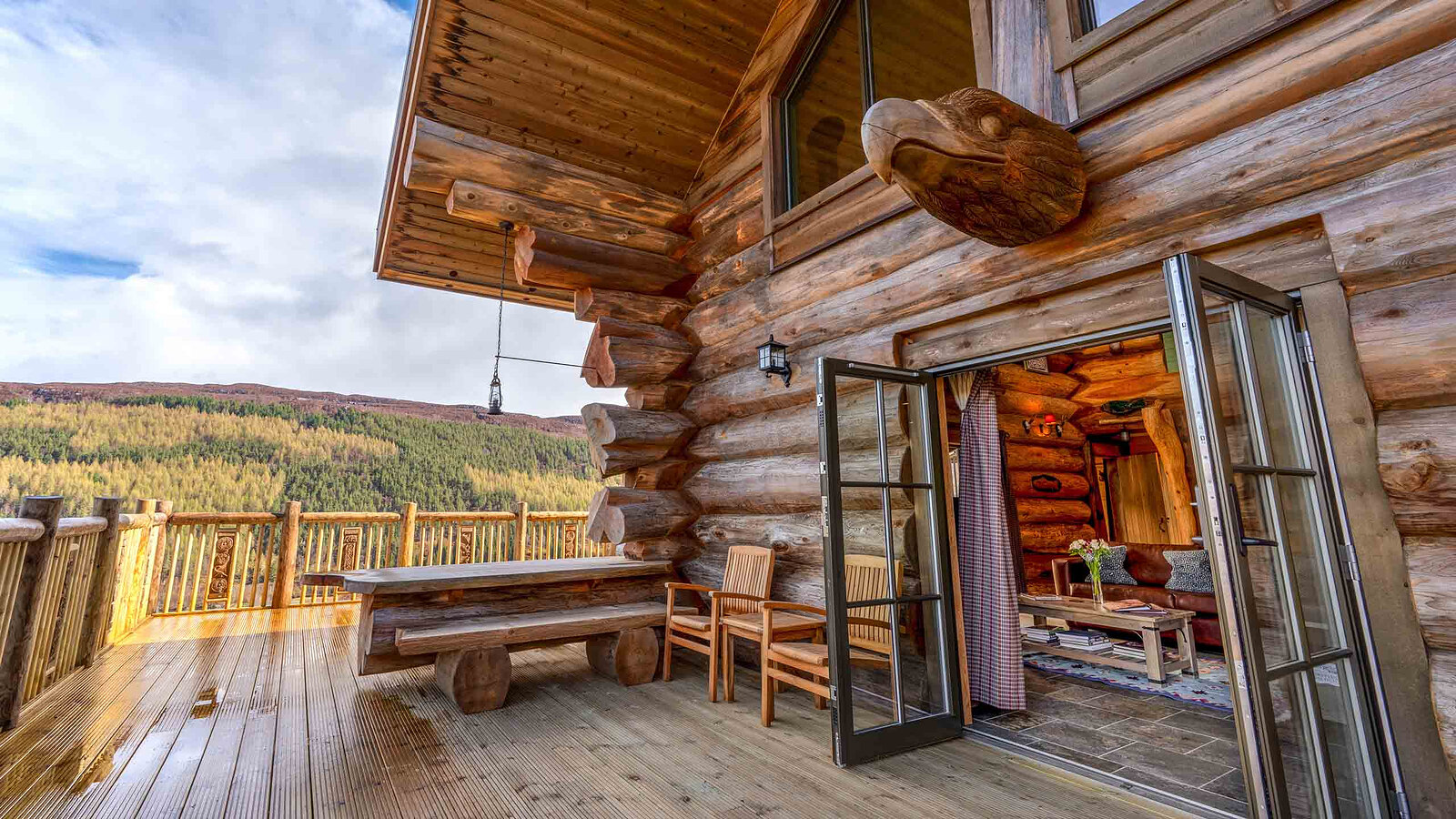 Aquila's covered deck with a log bench and table, its glass doors open to the living room, a carved eagle on the logs and the glen beyond.