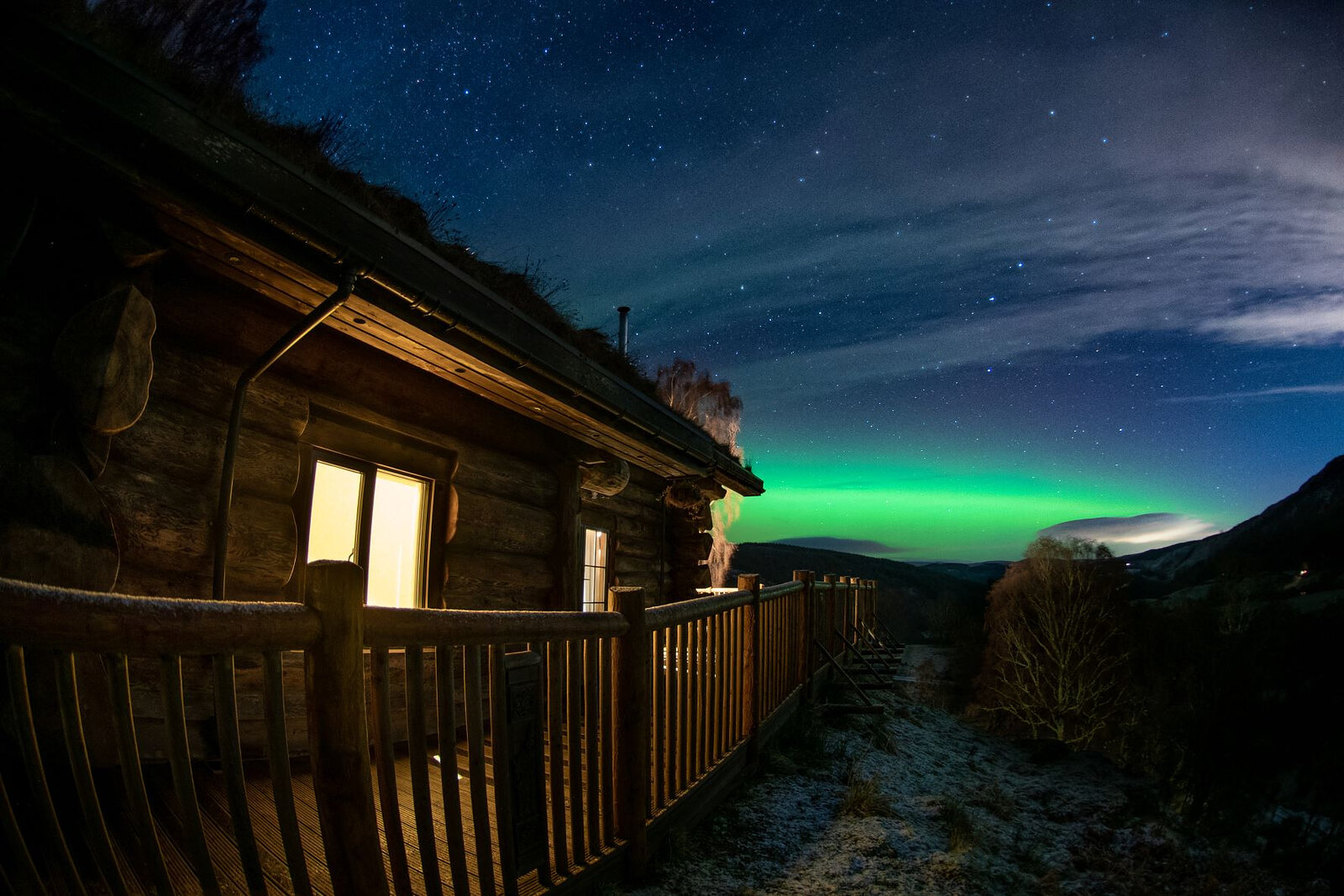The Northern Lights green over the glen beyond Aquila's lit windows and deck rail on a starry night.