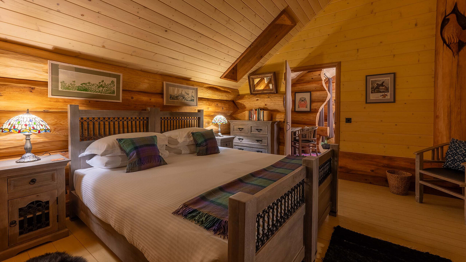 A double bedroom in Aquila under the sloping boards, a carved bed with tartan throws and a stained-glass lamp.