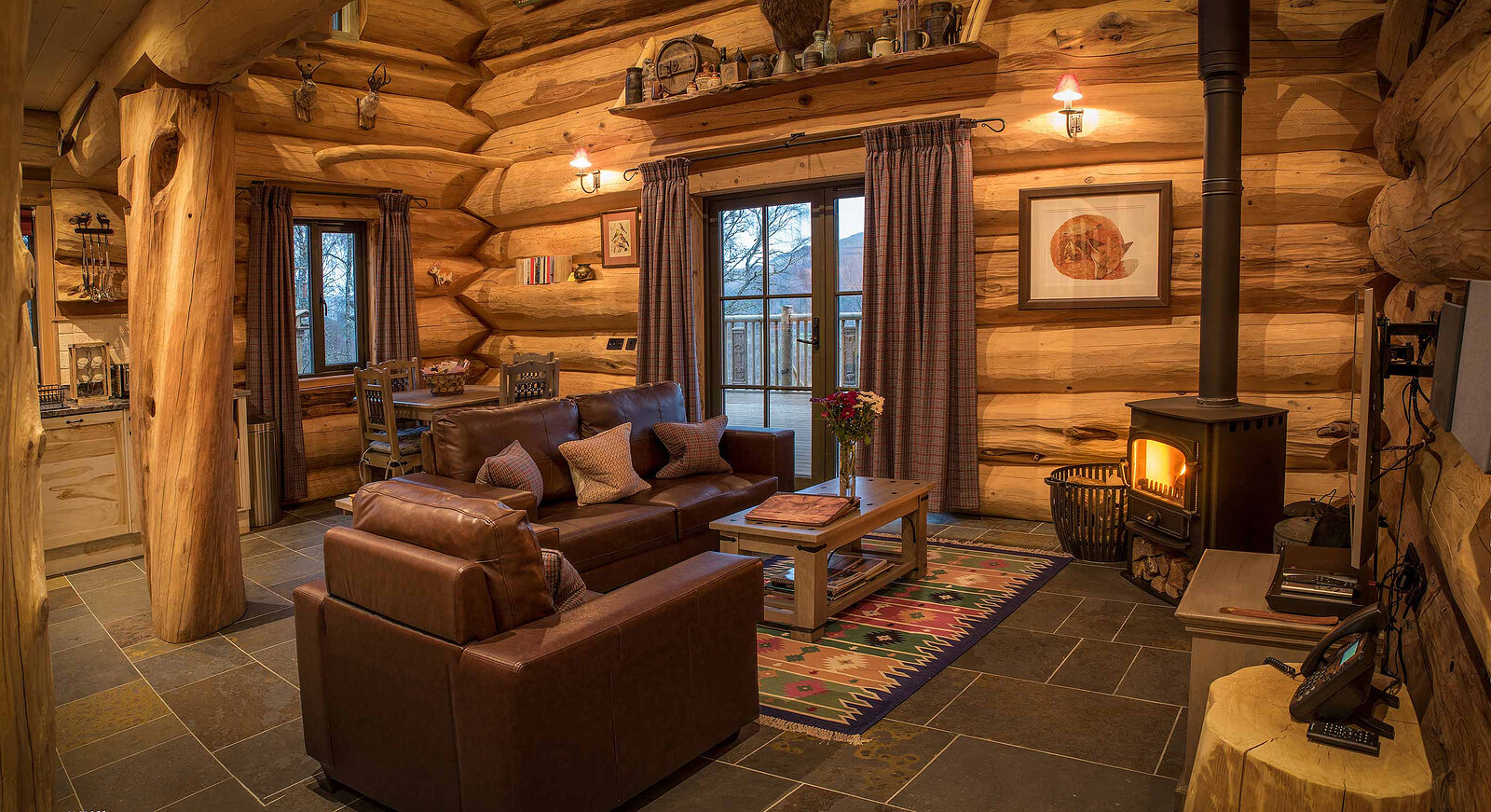 Ardea's living room: leather sofas round a table on a Himalayan rug, the stove lit by the glass doors and carved burr posts.