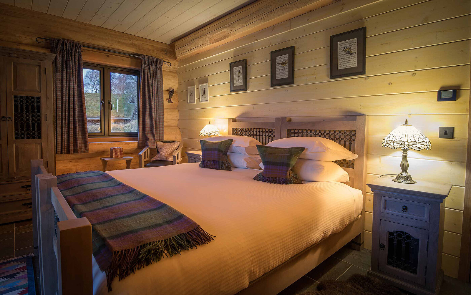 Ardea's bedroom: a carved bed with tartan throws, stained-glass lamps either side and bird prints on the pale boards.