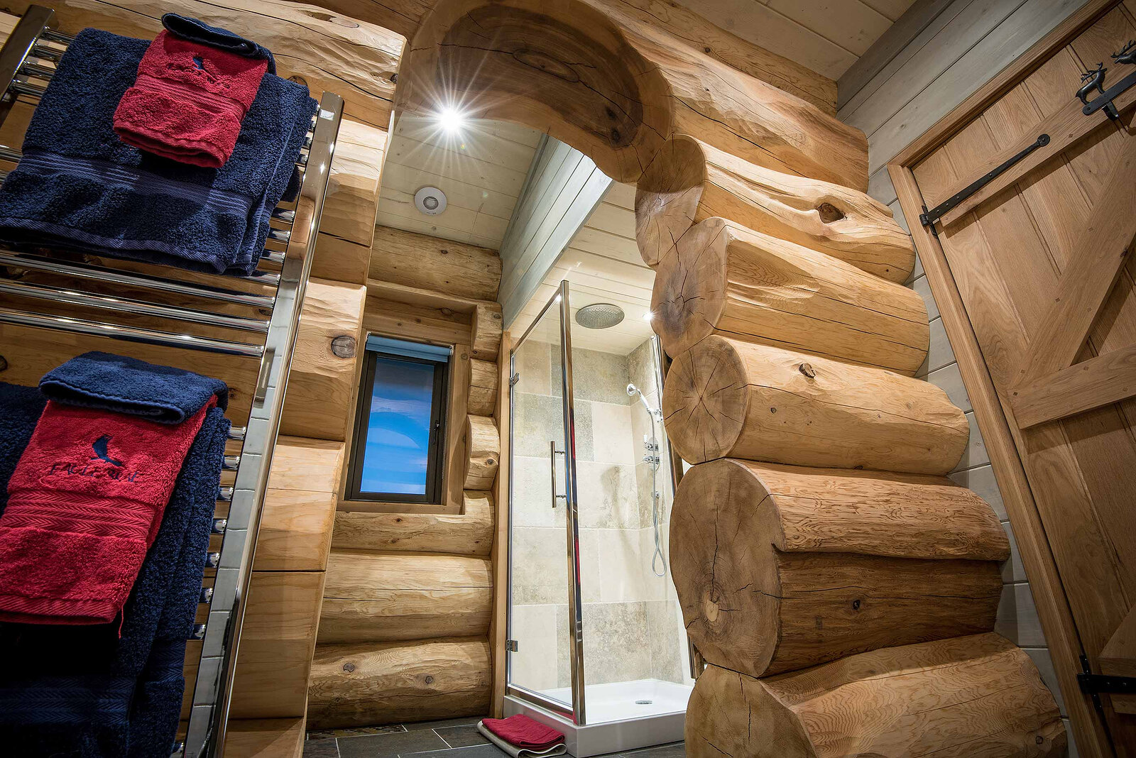 Ardea's bathroom through a log arch: red and navy towels on the rail and a glass shower.