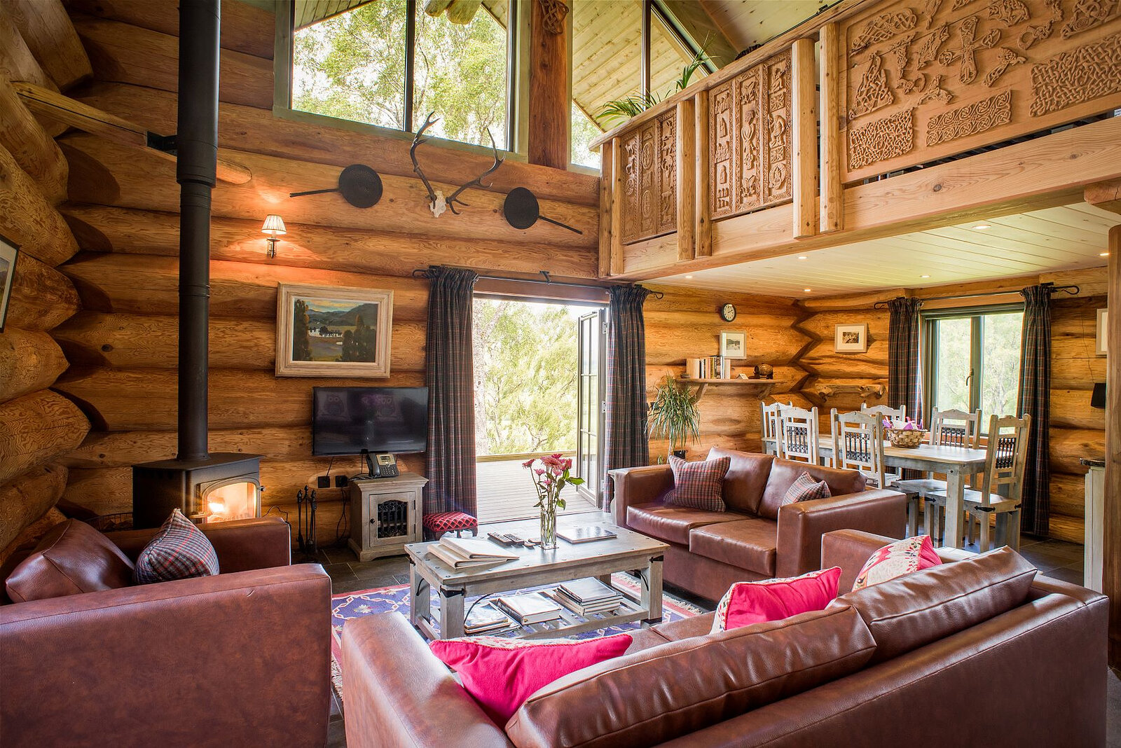 Buteo's living room: red leather sofas and pink cushions round the stove, the dining table by the open doors, a carved gallery above.