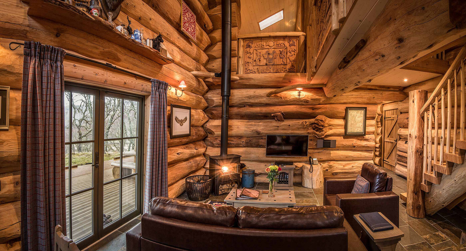 Certhia's living room: leather sofas round the stove, glass doors with tartan curtains to the deck and a log stair.