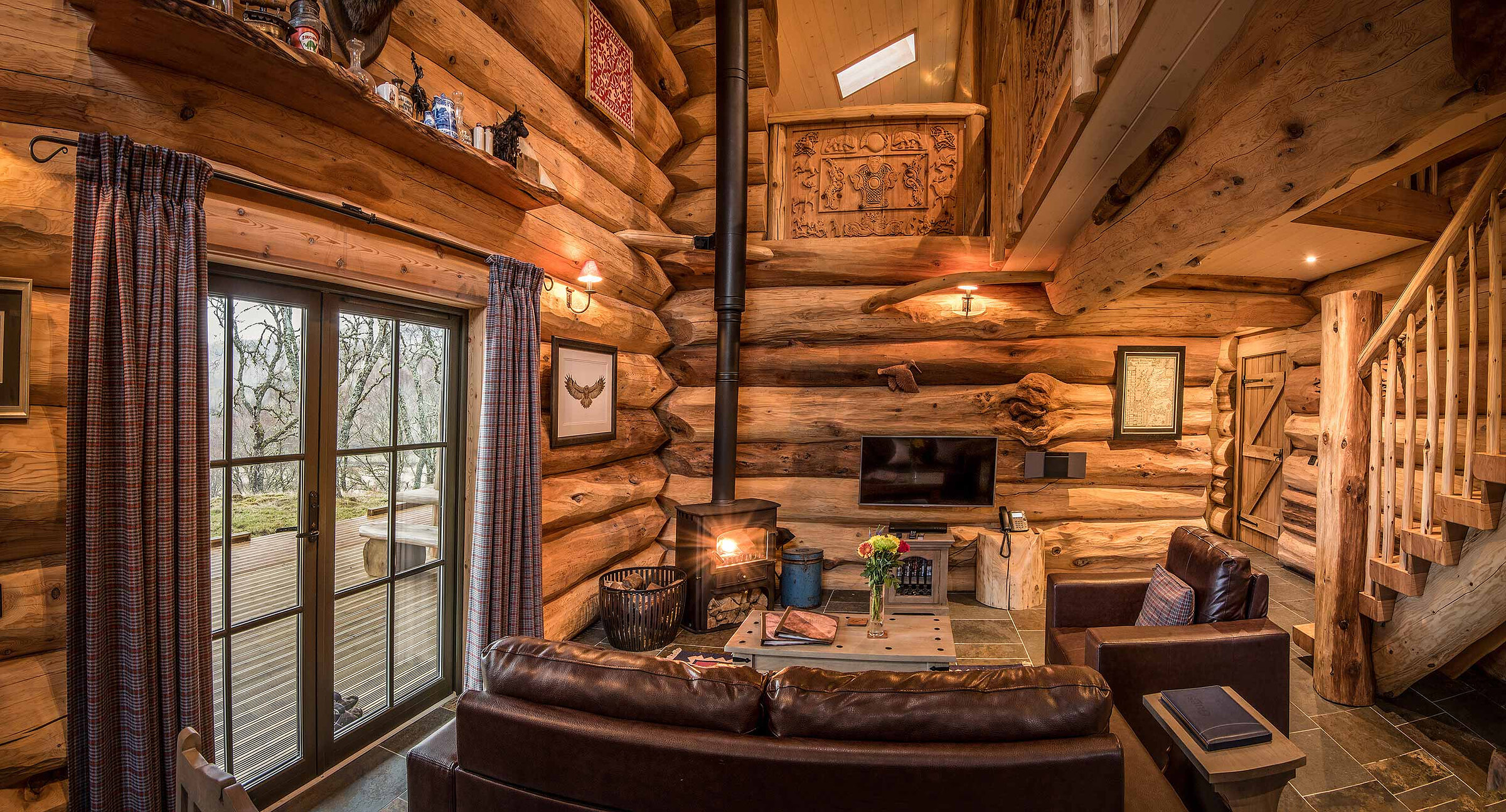 Certhia's living room: leather sofas round the stove, glass doors with tartan curtains to the deck and a log stair.