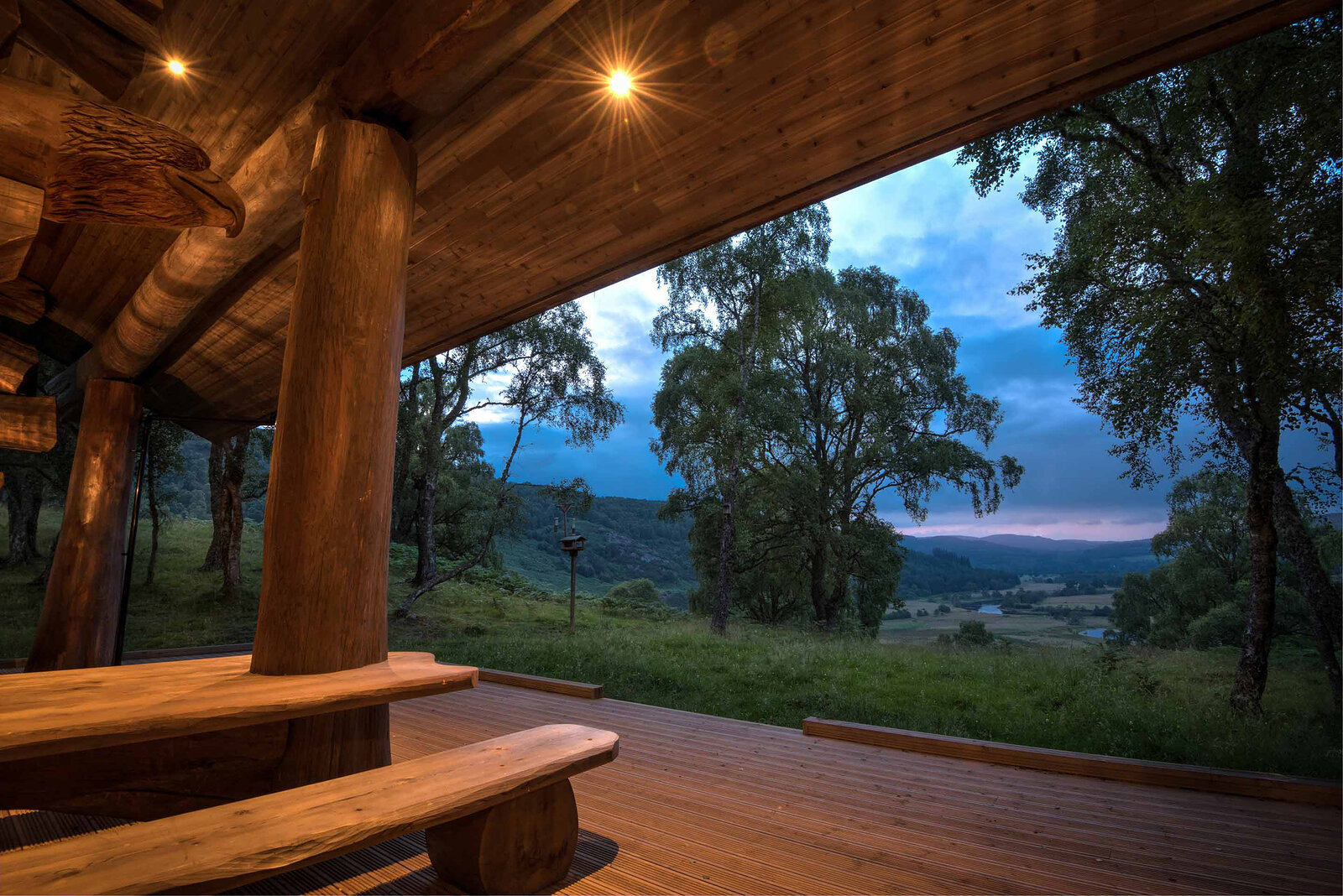 Certhia's covered deck at dusk, a log bench under the carved eagle and the glen blue beyond the birches.