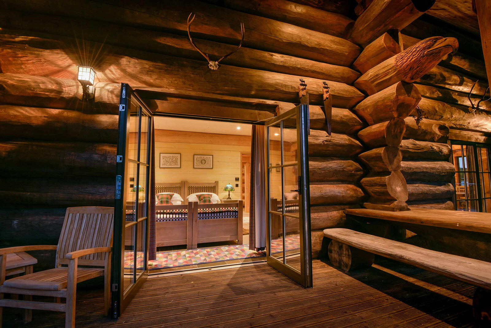 Cinclus's deck at night, its glass doors open to the lit bedroom, a carved eagle on the log corner.