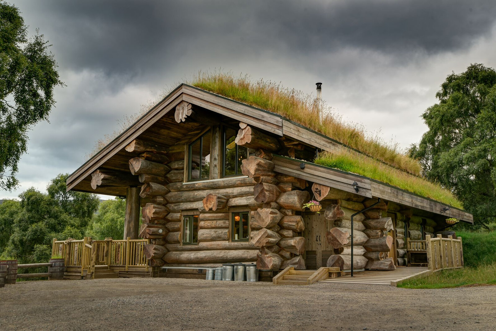 Cinclus under a grey sky: a turf-roofed log cabin with notched corners and a railed deck on its gravel yard.
