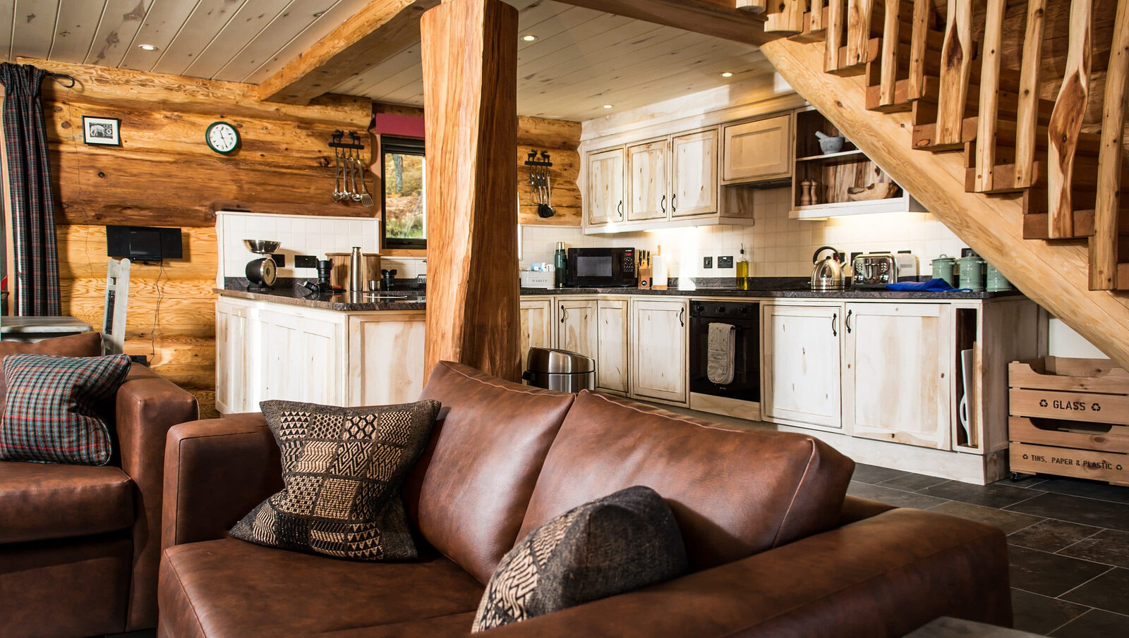 A cabin kitchen of cream cupboards under the stair, a leather sofa with patterned cushions in front.