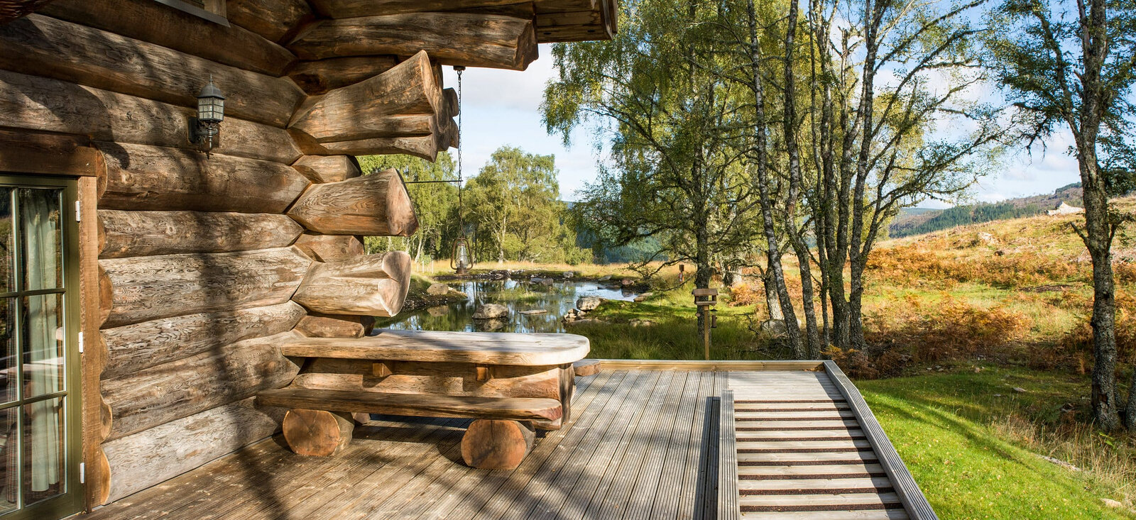 A log table on Loxia's deck in the sun, a ramp down to the grass and the pond through the birches.