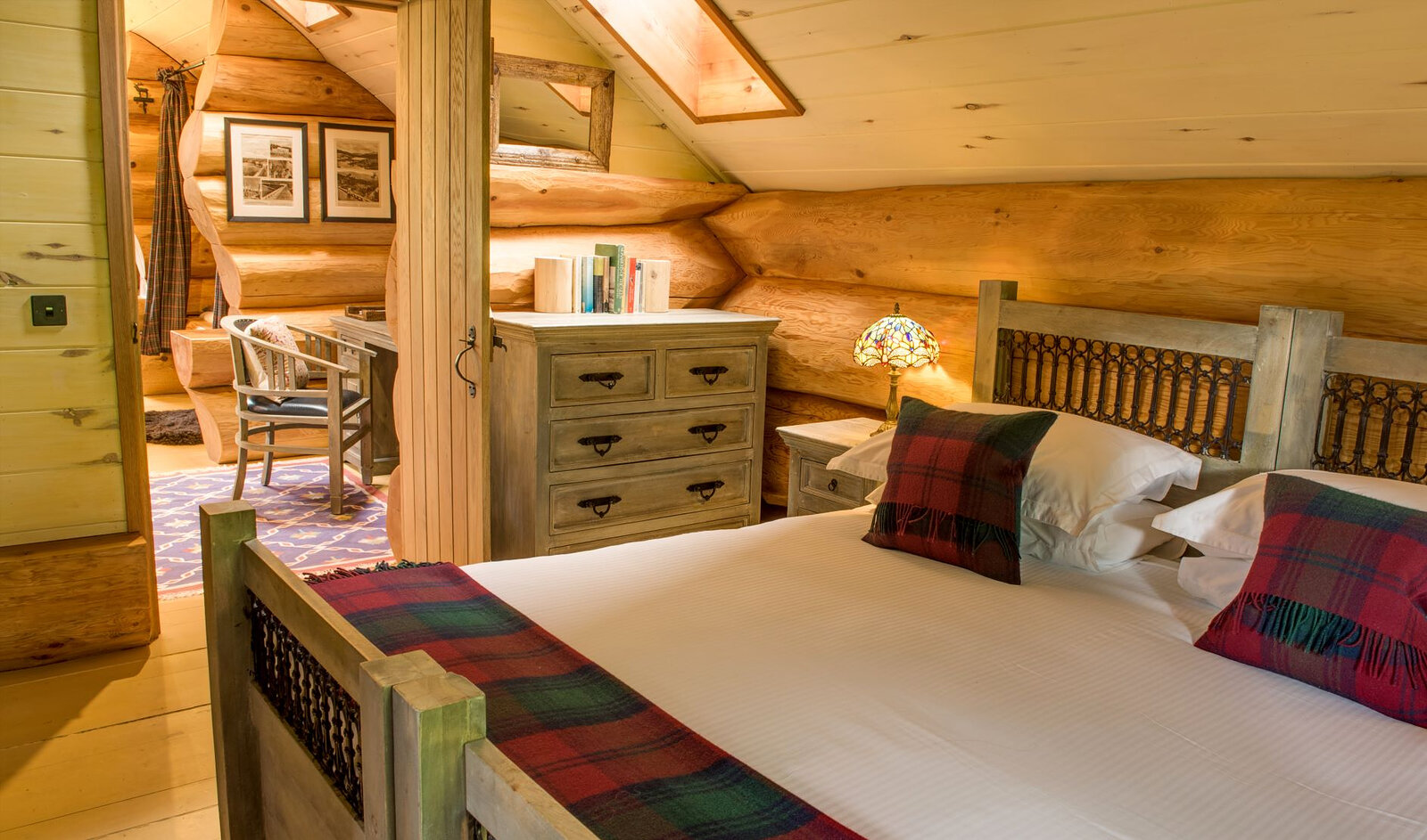 A bedroom under Loxia's sloping roof: a carved bed with red tartan cushions, a chest of drawers and a desk by the post.