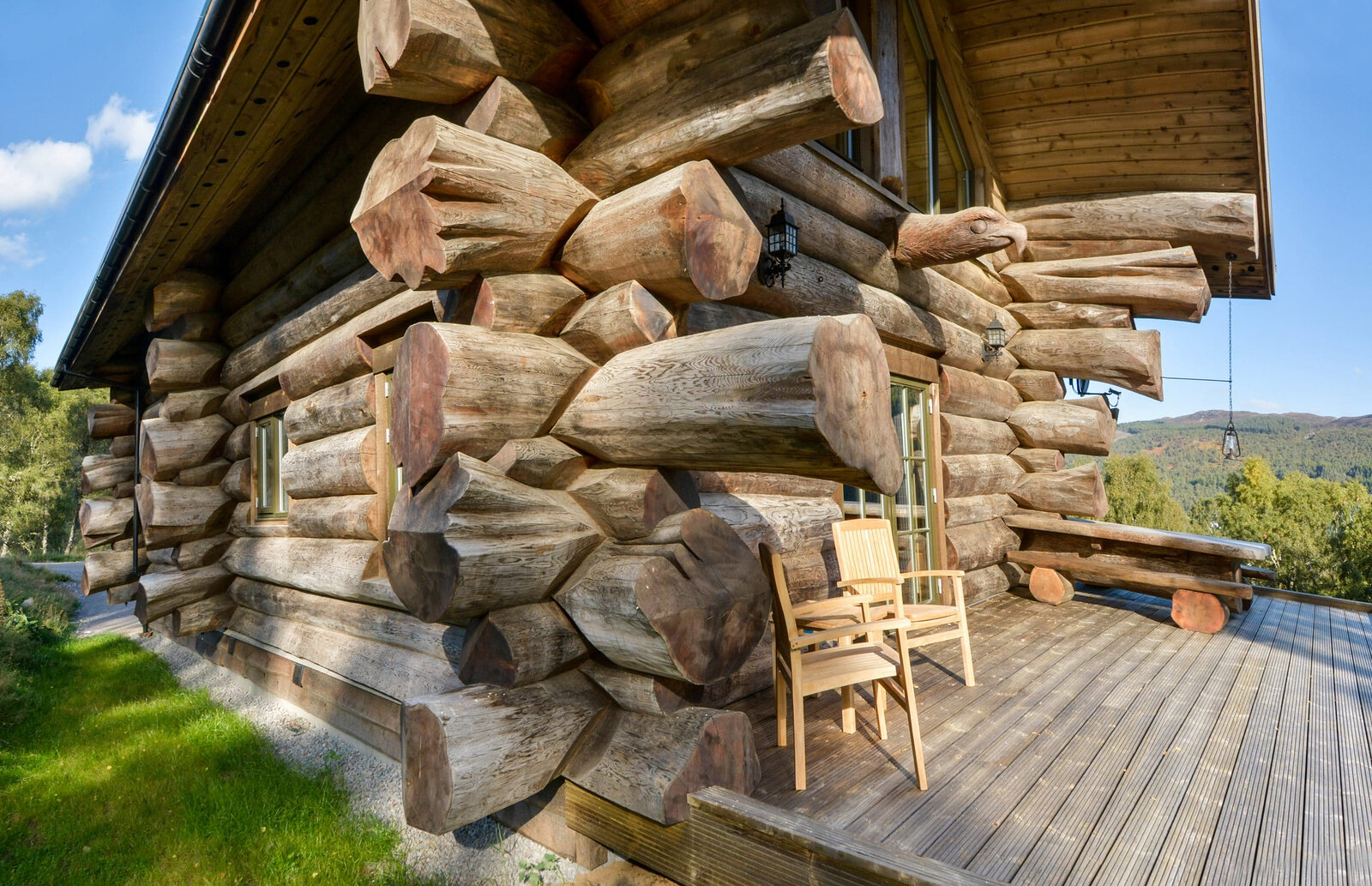 The great notched log corner of a cabin over a sunny deck, a chair and log bench by the wall and the glen beyond.