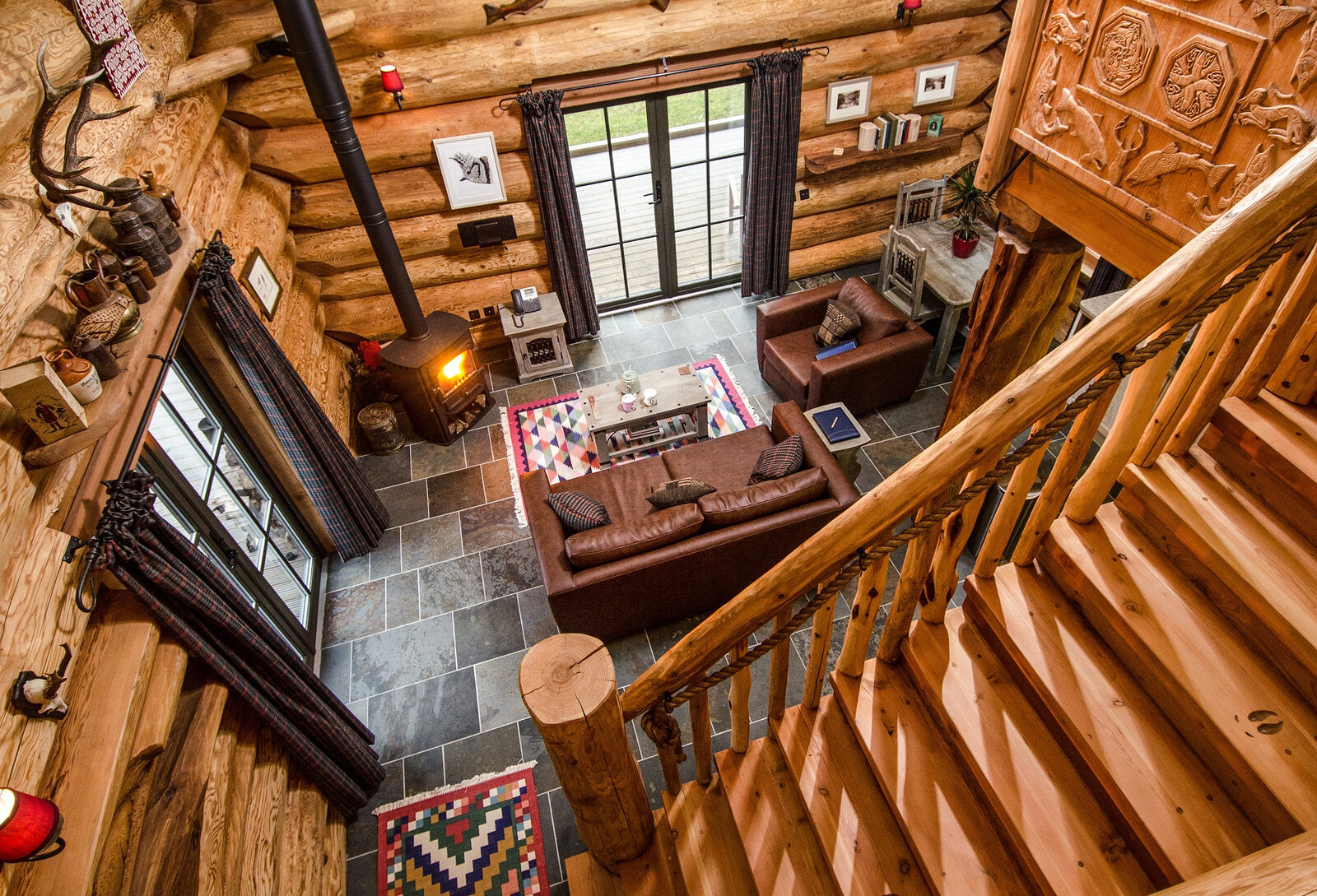 Down the log stair into a cabin's living room, a leather sofa on a bright rug by the lit stove.