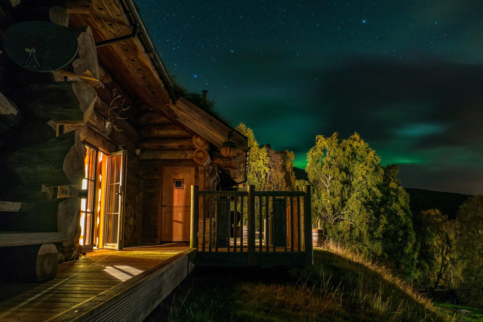 A cabin's deck at night, the door lit and the Northern Lights faint over the birches.