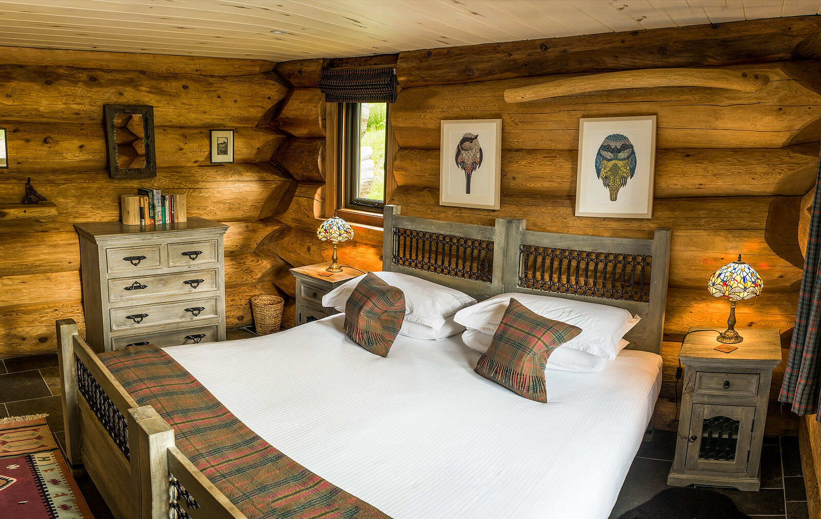 Parus's bedroom: a carved bed with tartan cushions under framed tit prints, lamps glowing either side.