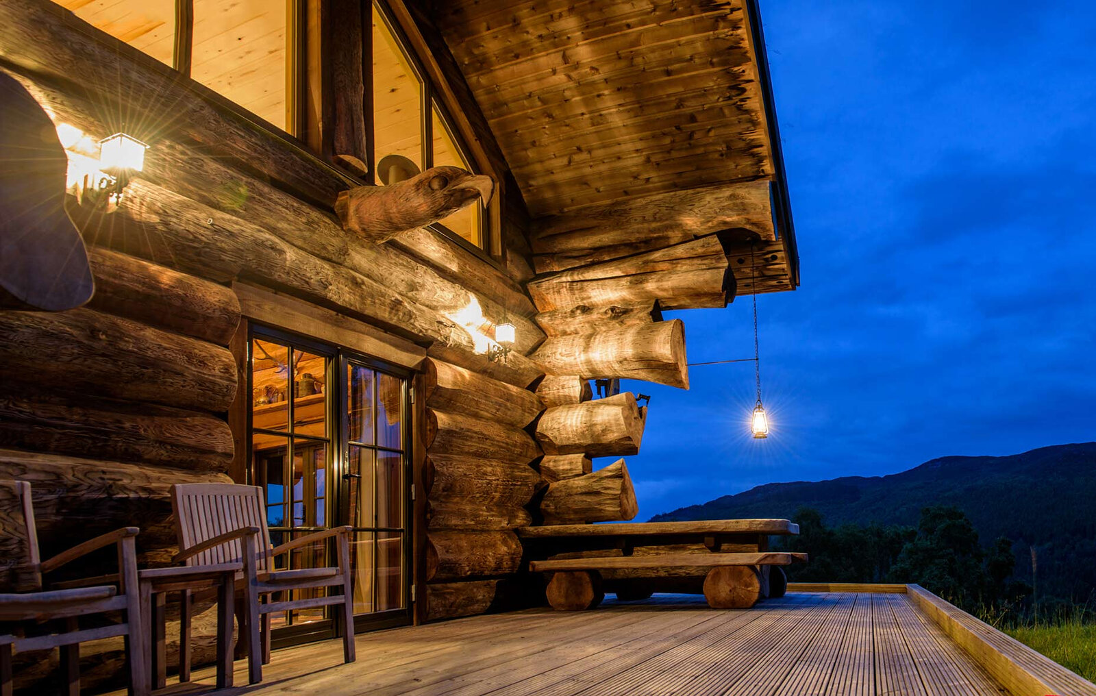 A cabin's deck at dusk, lanterns lit on the log walls and chairs by the glass doors, the hills dark blue.