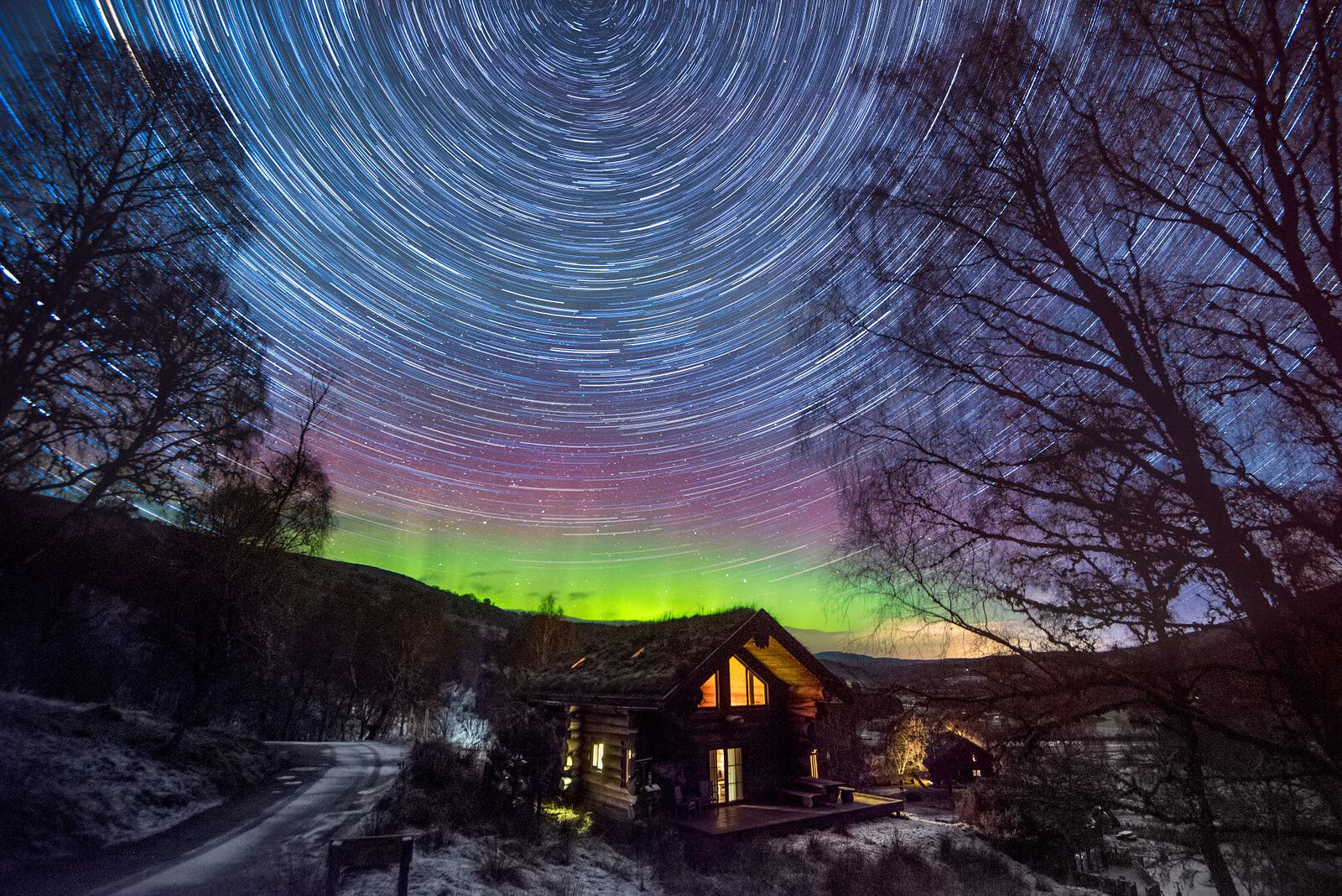 Star trails circling over a cabin lit among bare trees, the Northern Lights green on the horizon.