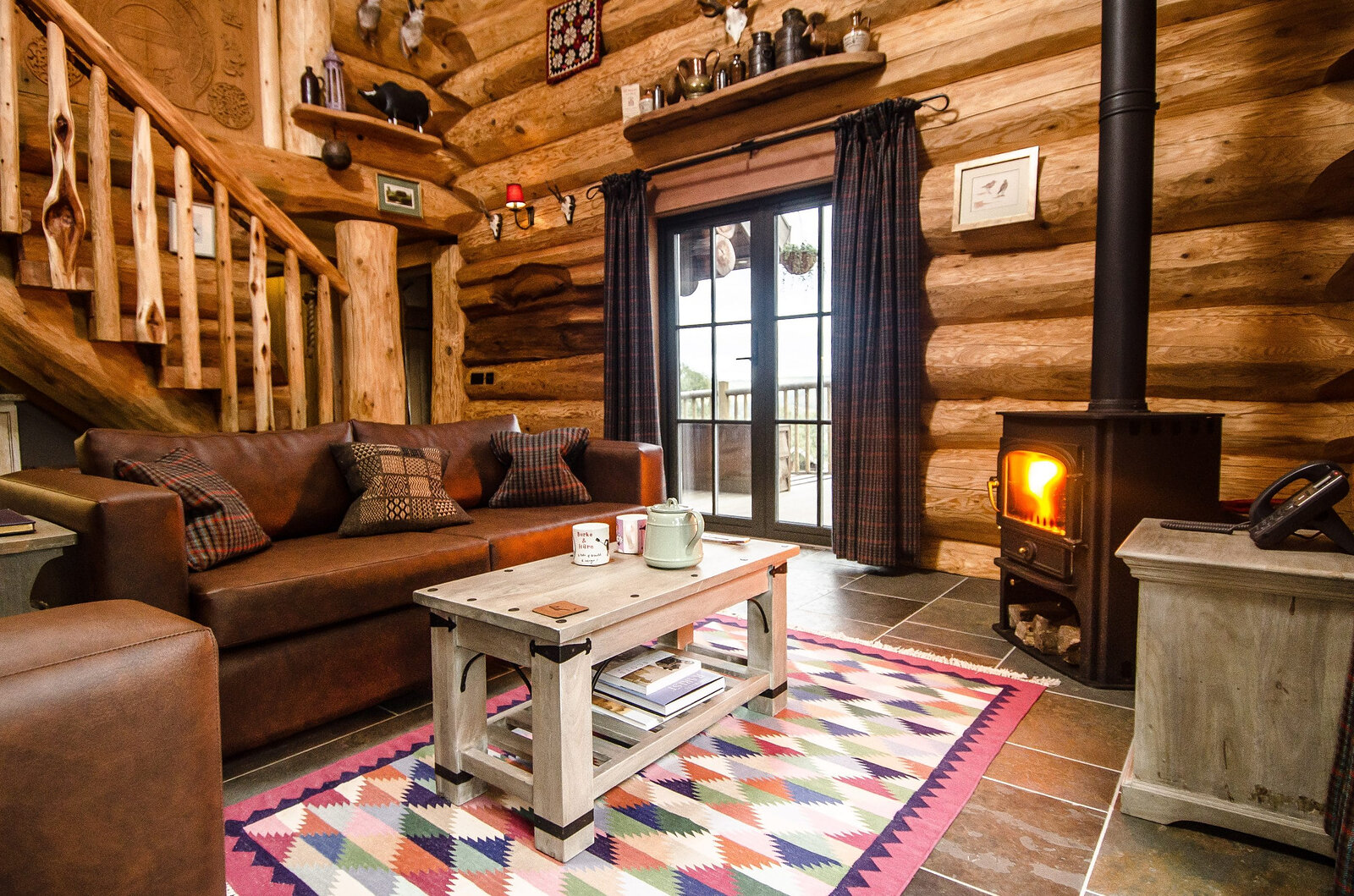 Parus's living room: a leather sofa and a patterned rug by the lit stove, the log stair and glass doors beside.