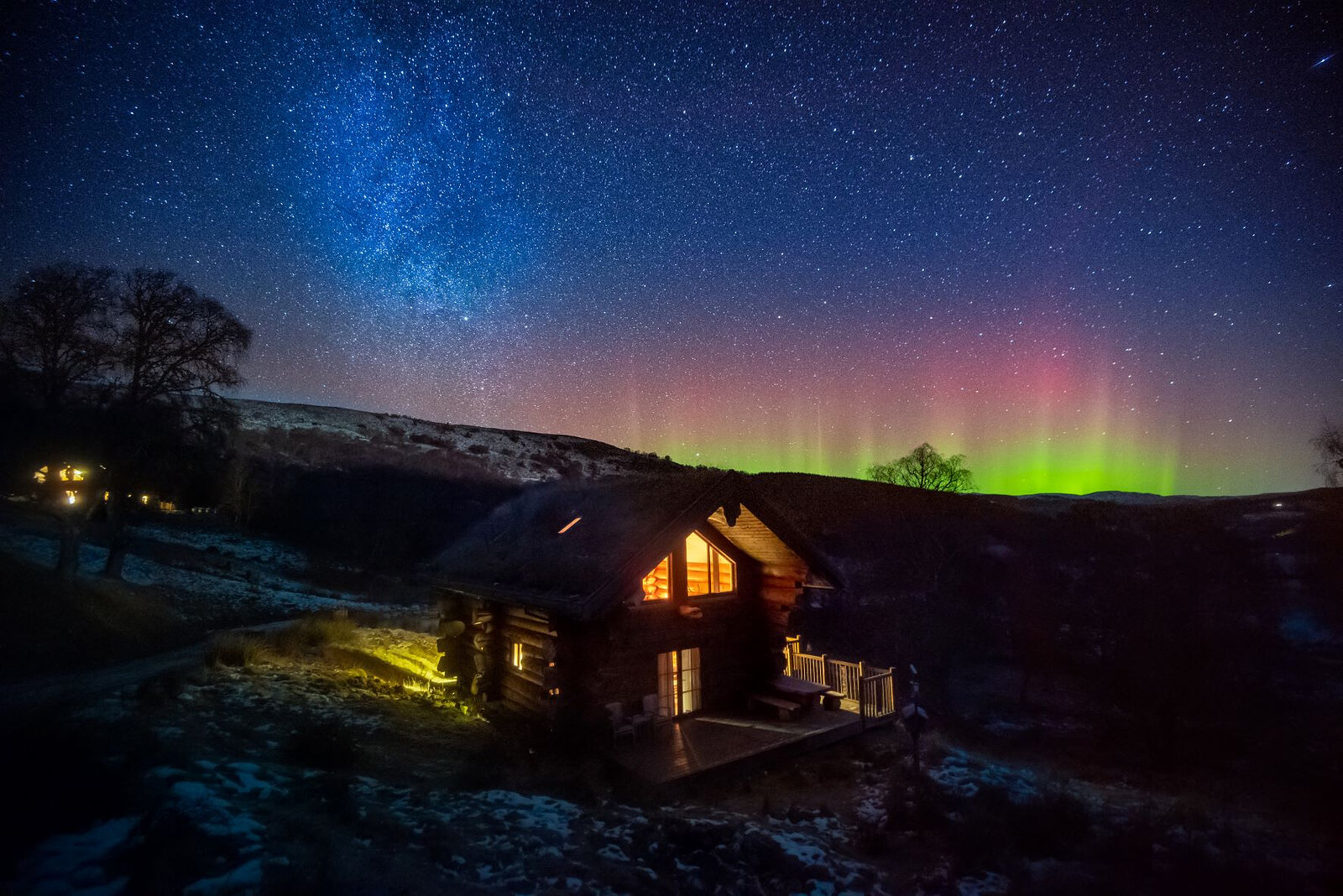 The Northern Lights red and green over the glen behind a lit cabin, the Milky Way above.