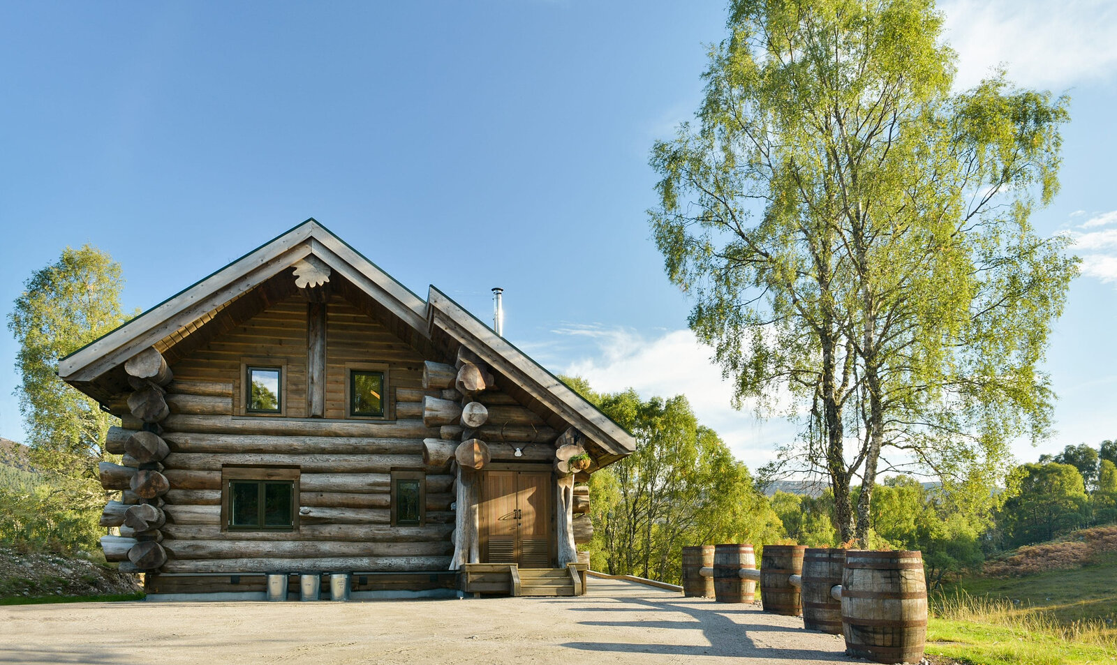 Sylvia in sunshine: a log cabin with a pale gable and oak door, whisky barrels along its gravel yard and a birch beside.
