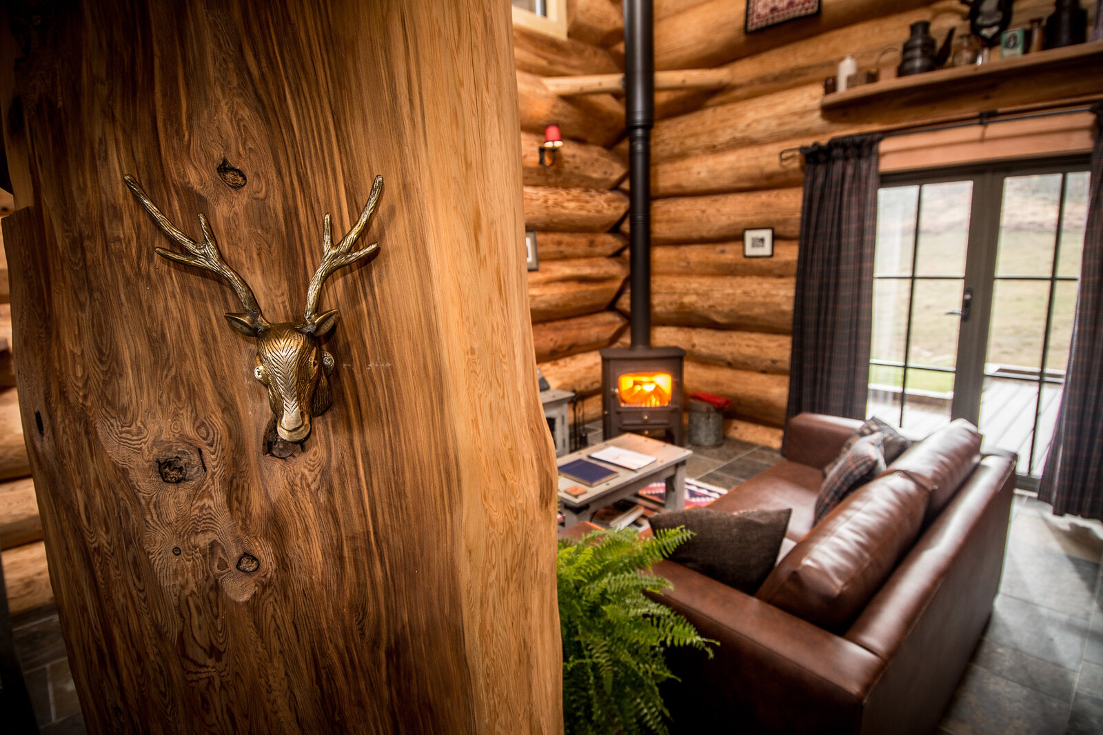 A brass stag's-head hook on a log post in Sylvia, the leather sofa and lit stove beyond.