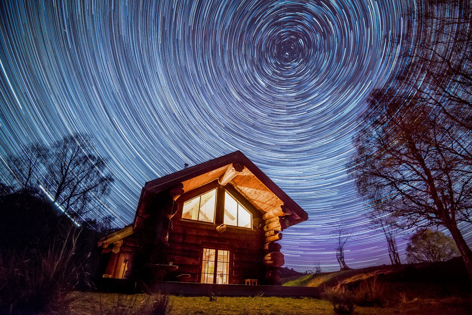 Star trails wheeling over Sylvia's lit gable at night, bare trees either side.