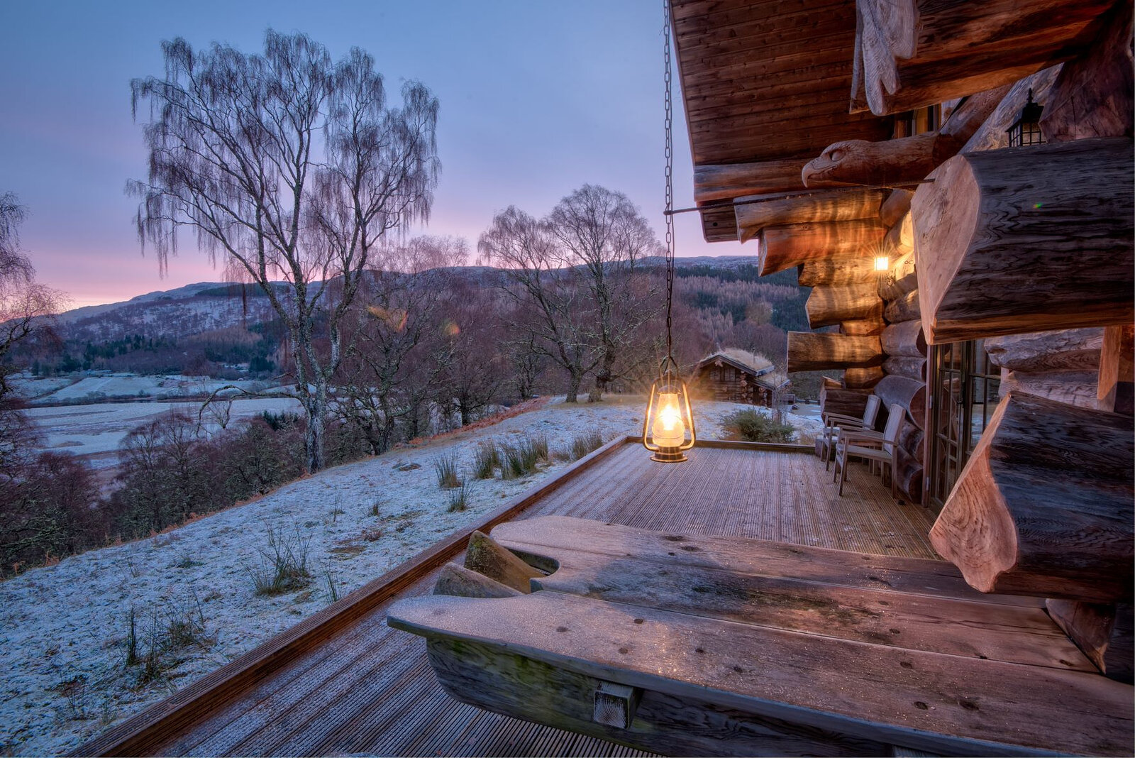 Tringa's deck at dawn in frost, a lantern lit over the log bench and the glen pink beyond the birches.