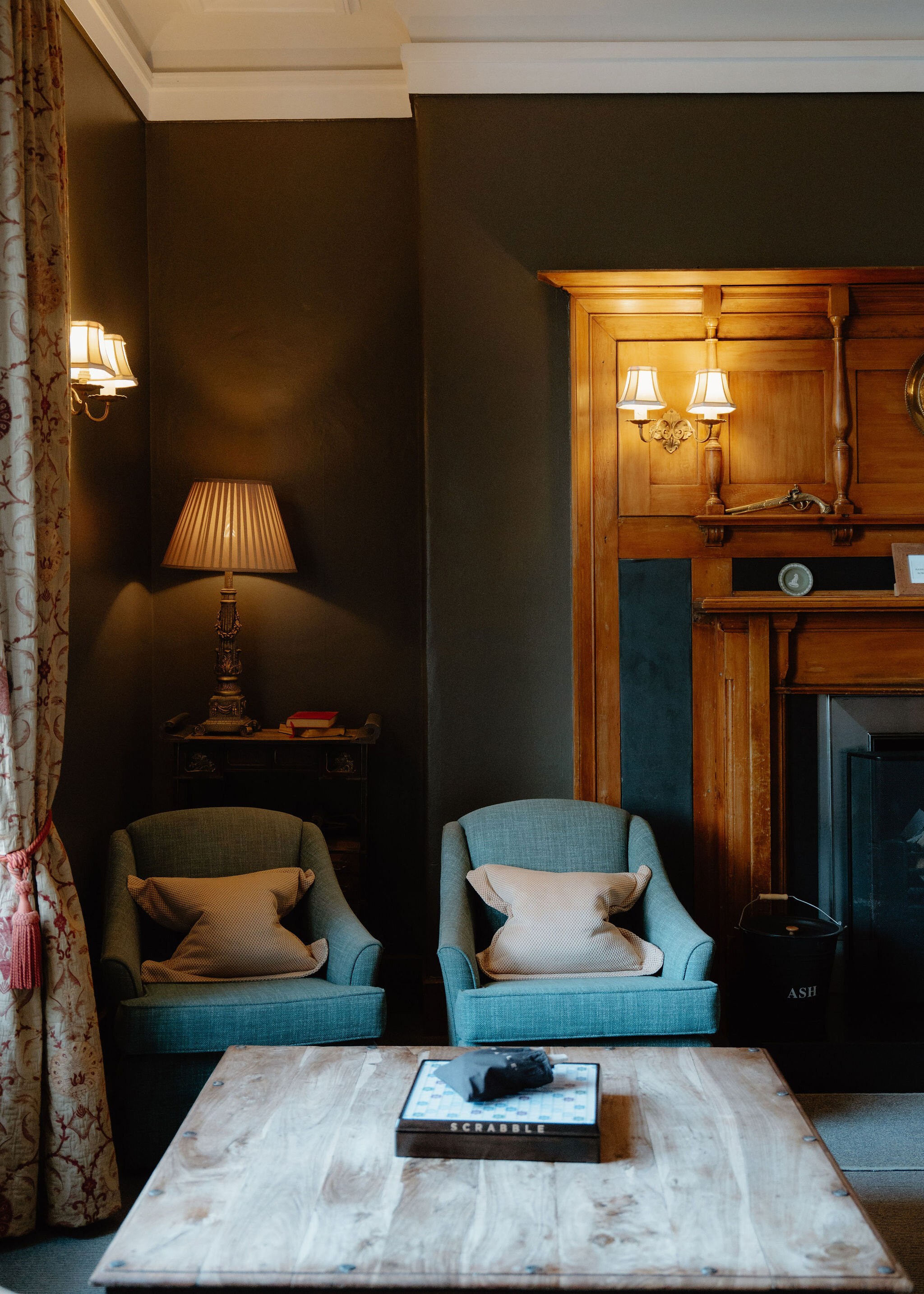 A dark green sitting room: two teal armchairs, a pale wooden coffee table with a Scrabble box, a lamp and a panelled fireplace.