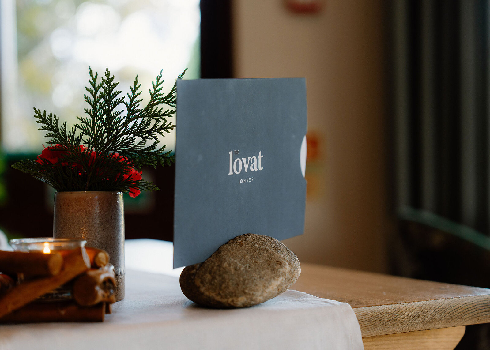 A landscape frame of the menu in its river stone, a candle in a holder of cut logs and evergreen with red flowers.