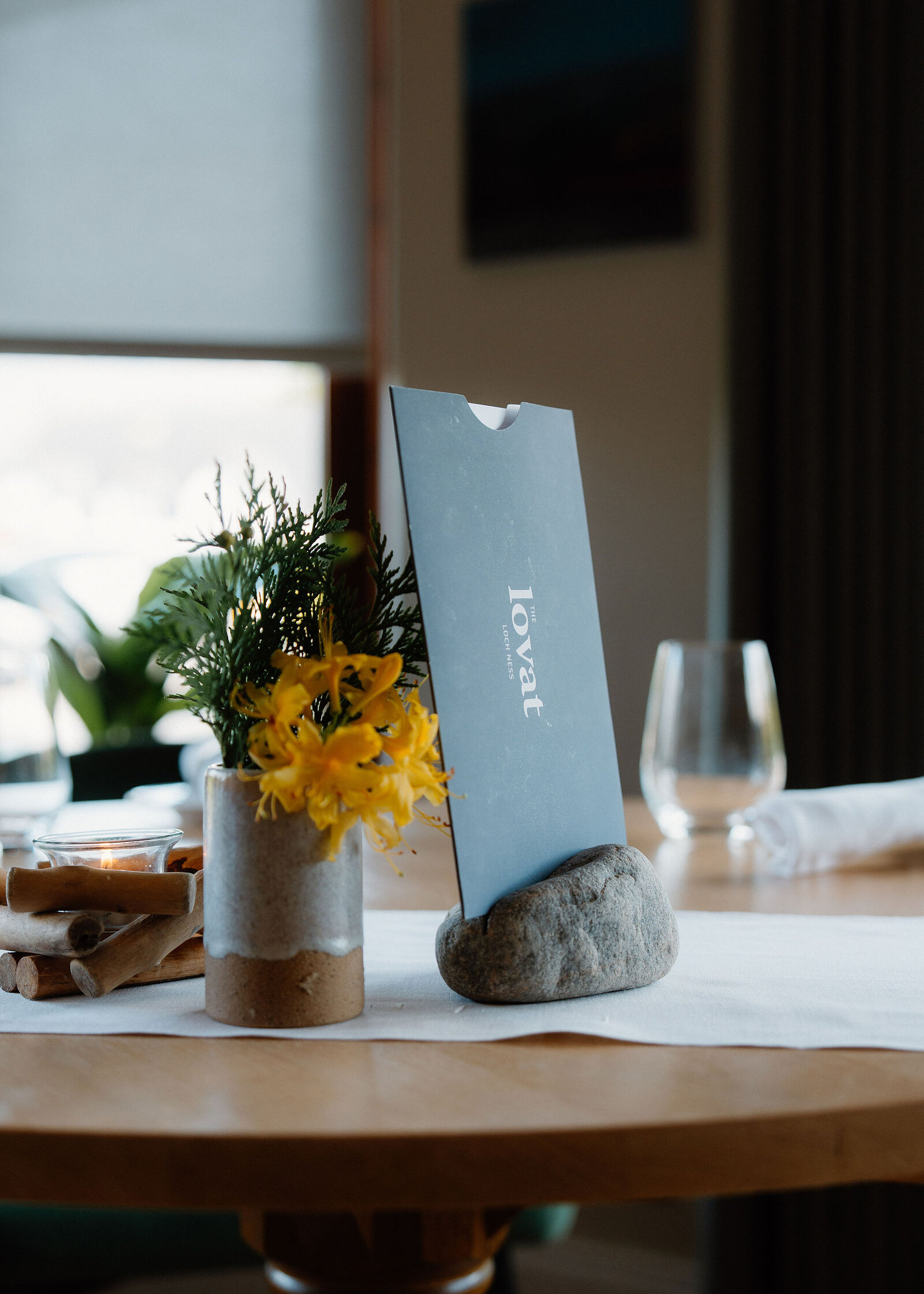 A round oak table laid with a linen runner, yellow flowers in a stoneware vase, the menu in its stone, glasses and napkins.
