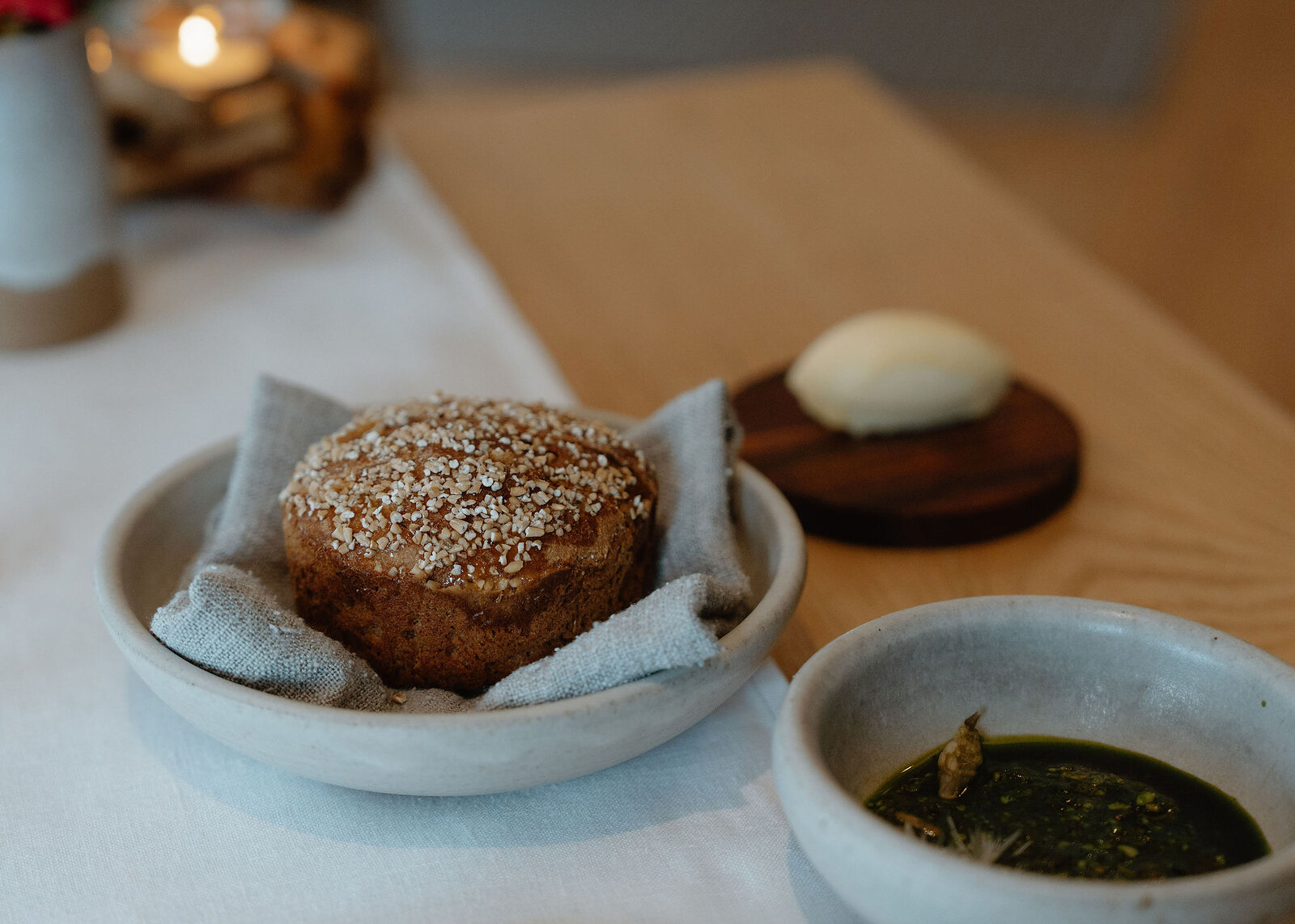 A landscape frame of the oat-crusted loaf in its bowl, with butter on a wooden round and a dish of green herb oil.