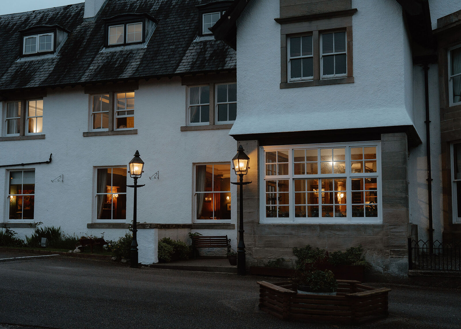 The Lovat at dusk: white front, slate roof and dormers, lit lamps and the glowing bay window, a planter on the drive.