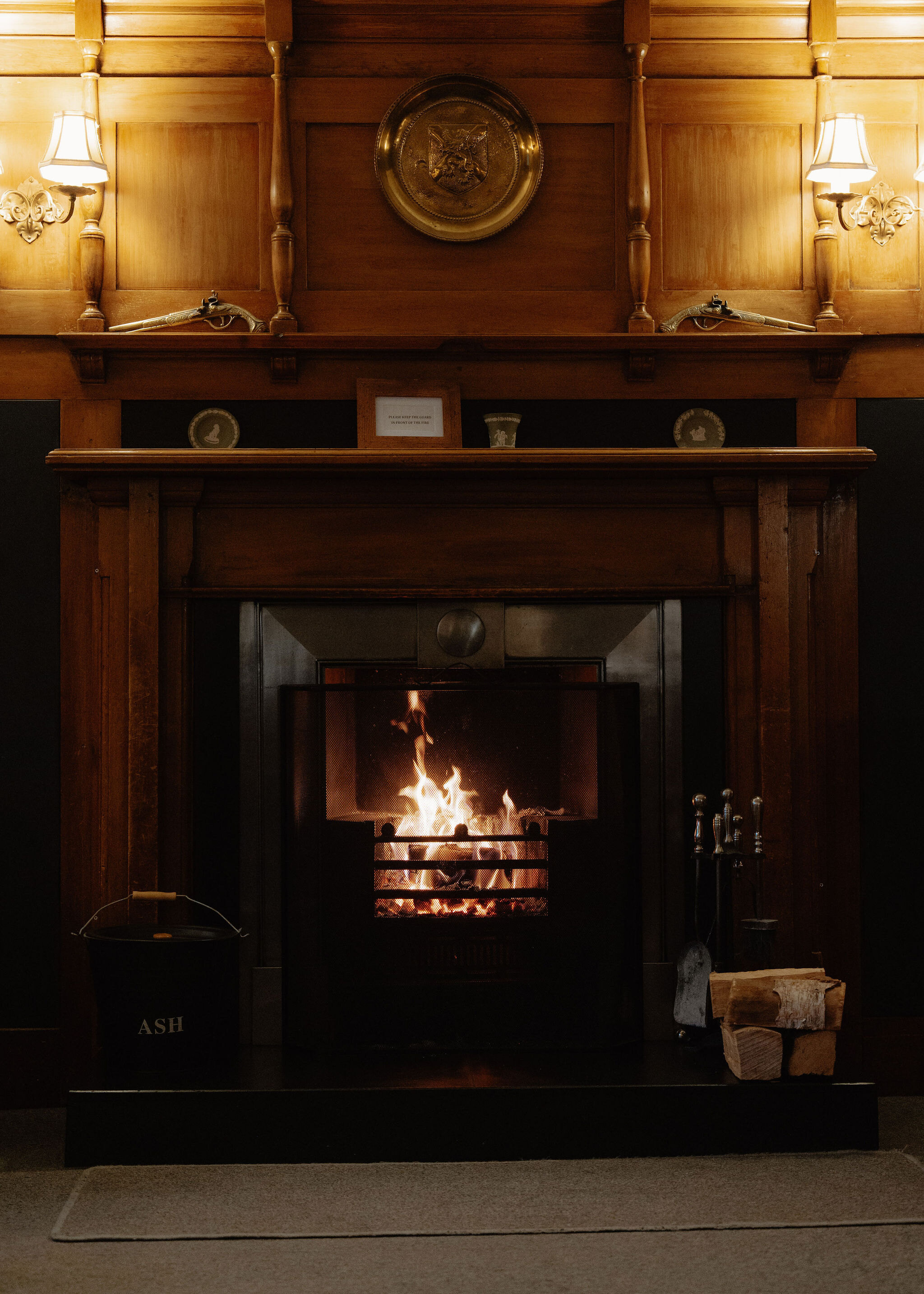 A log fire burning in a cast-iron grate under a panelled wooden mantel, twin wall lights, a brass plaque and an ash bucket.