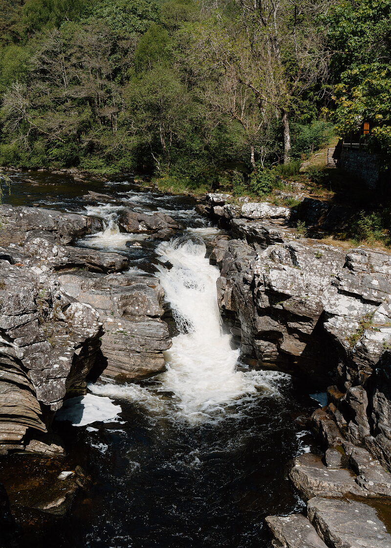 Looking down on a waterfall pouring white through a channel in grey rock into a dark pool, woods above.