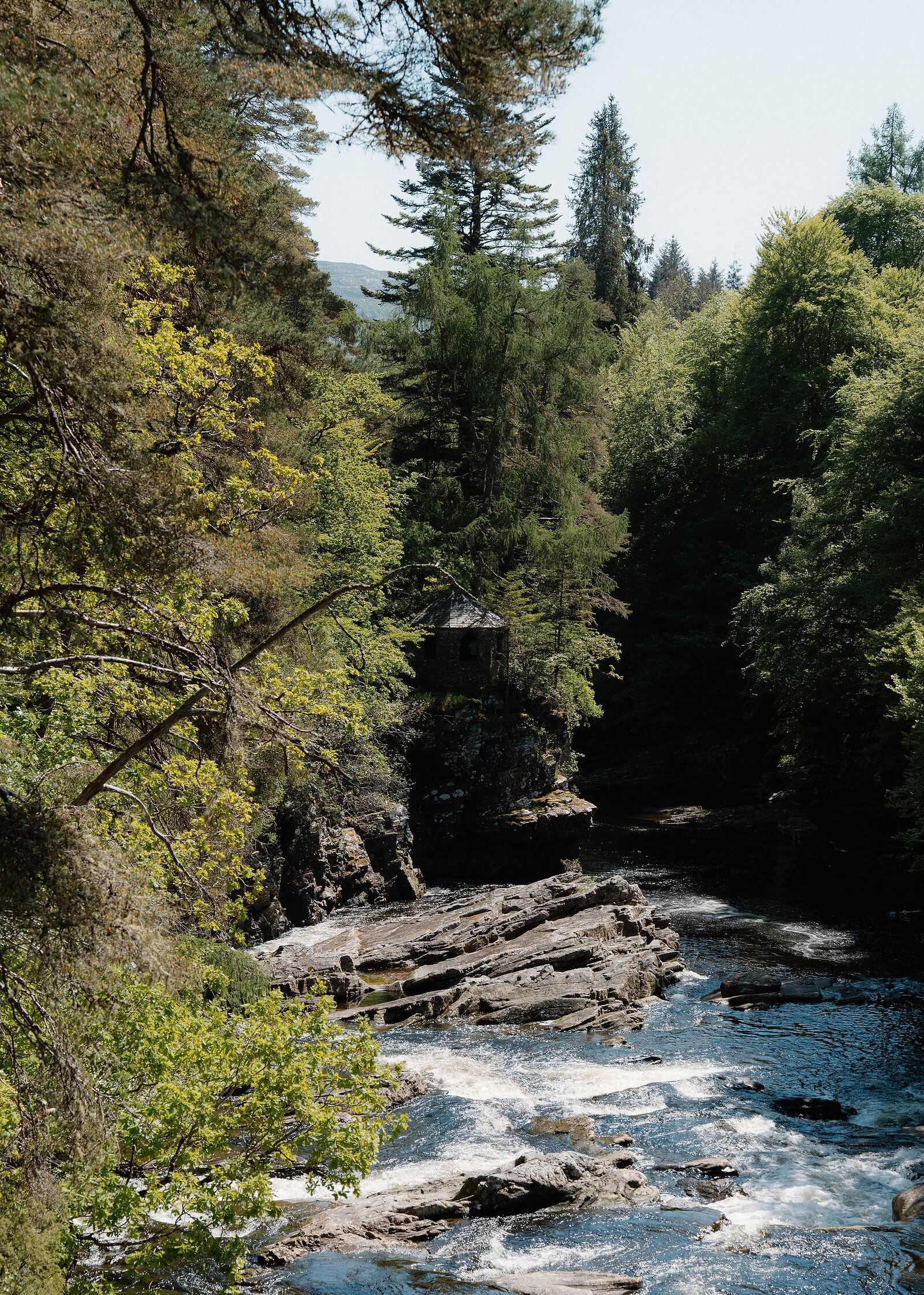 The river running white over rock ledges in a wooded gorge, a small stone summerhouse on the far bank.