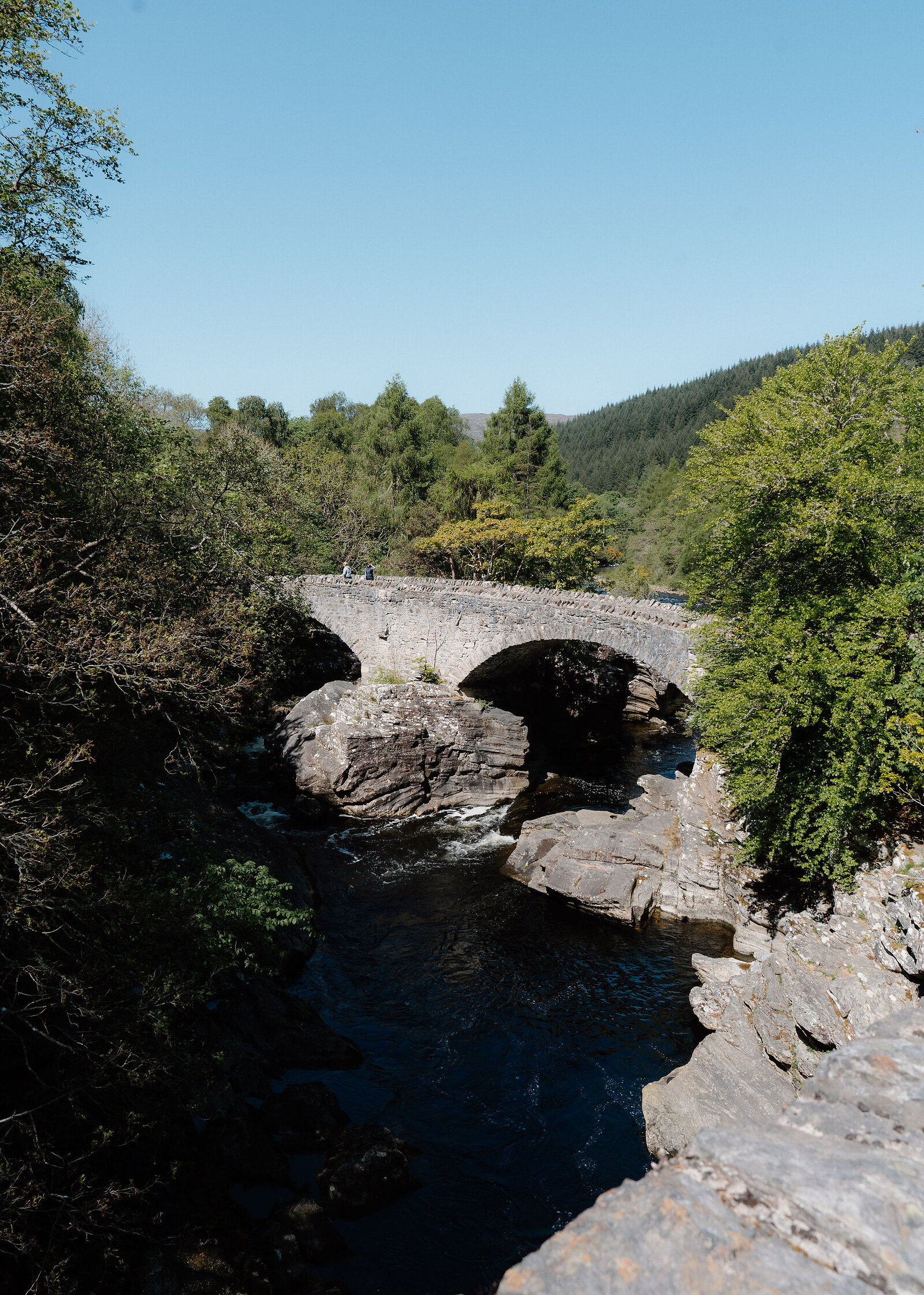 A twin-arched stone road bridge over a dark river pool and rocks, forested hills under a clear blue sky.